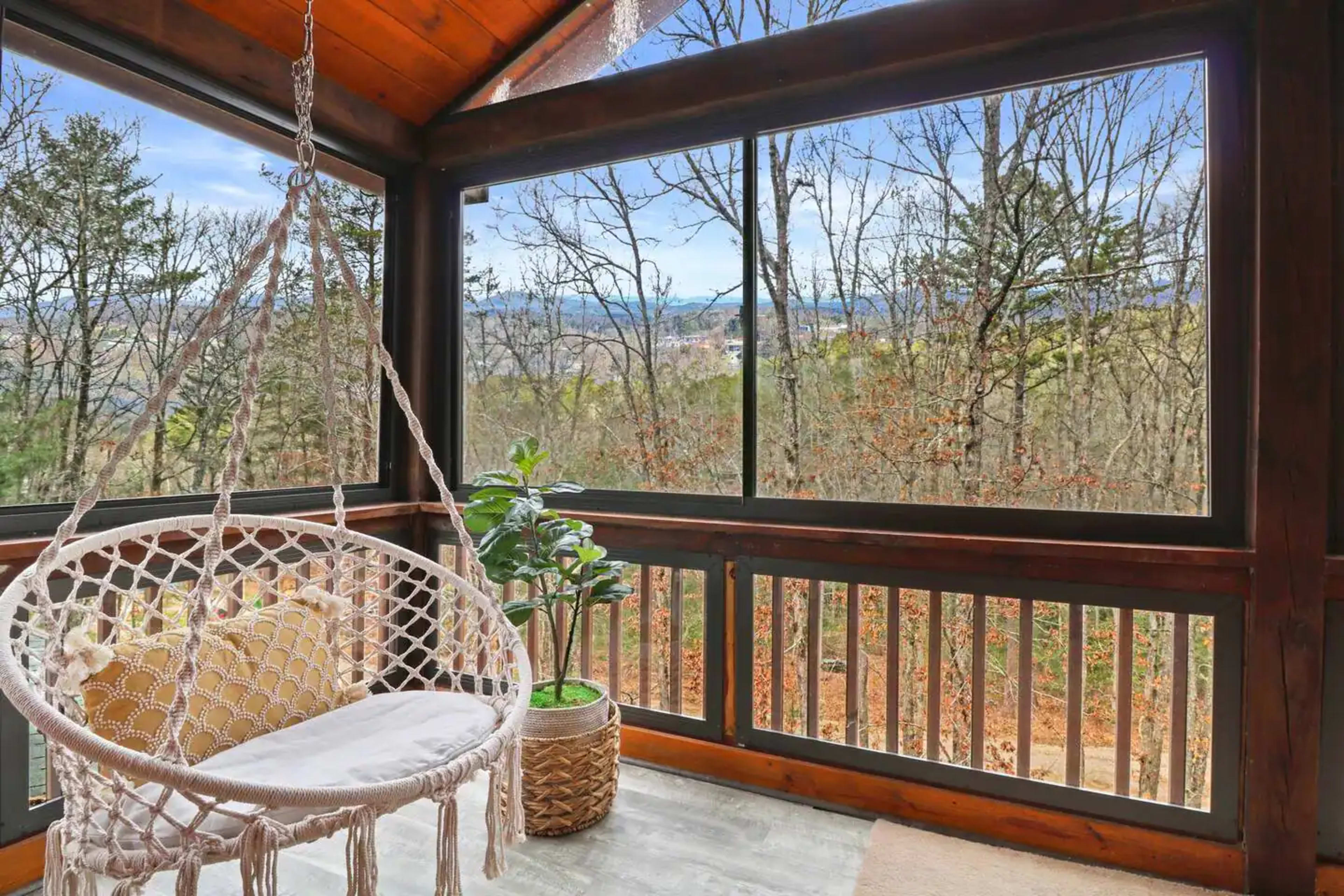 A cozy screened-in porch features a hanging macramé chair and a potted plant with a view of the wooded landscape outside.