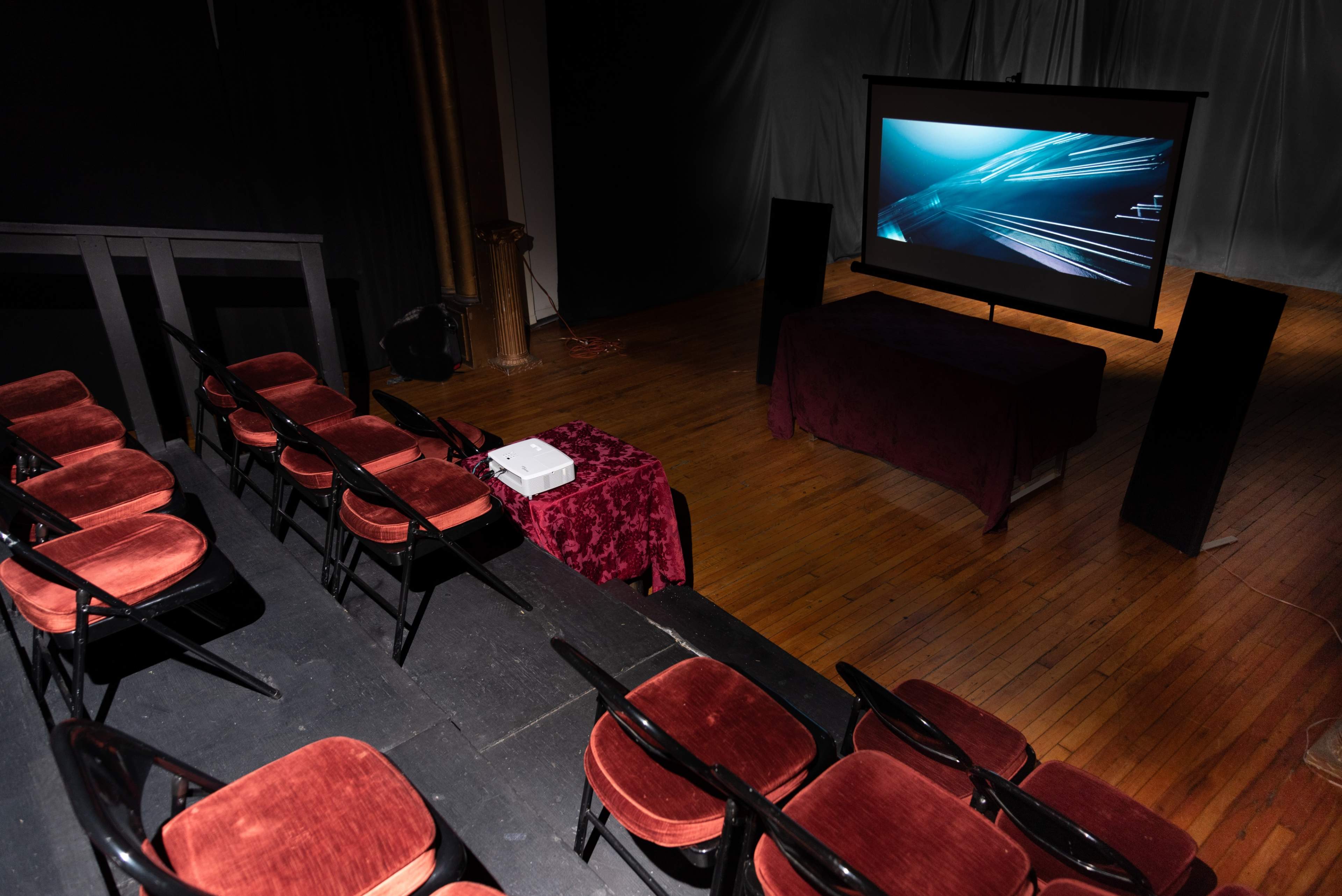 The image shows a theater stage setup with empty red chairs facing a large projection screen displaying abstract visuals.