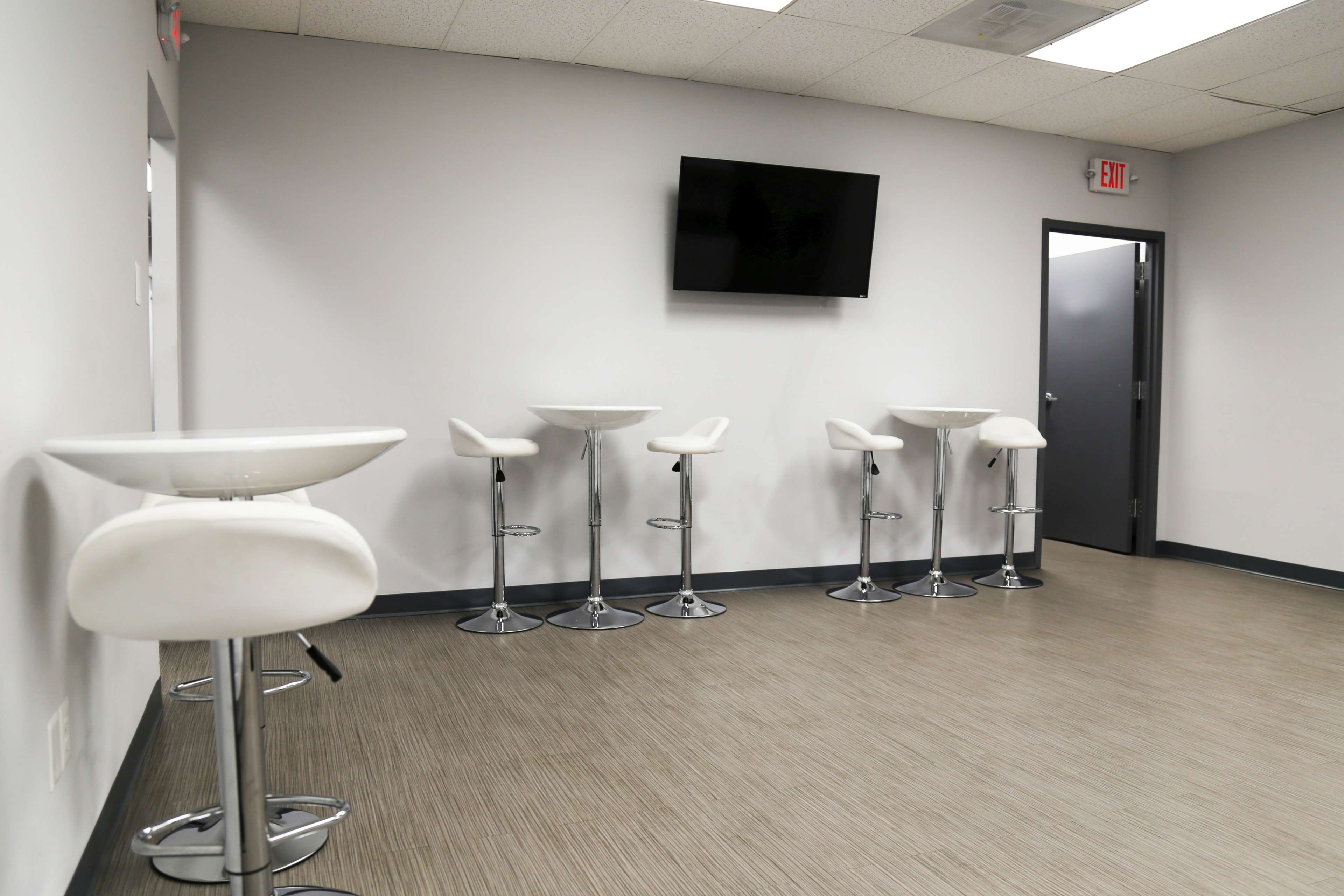 A plain room with four high stools and small tables against a light gray wall, featuring a wall-mounted television.