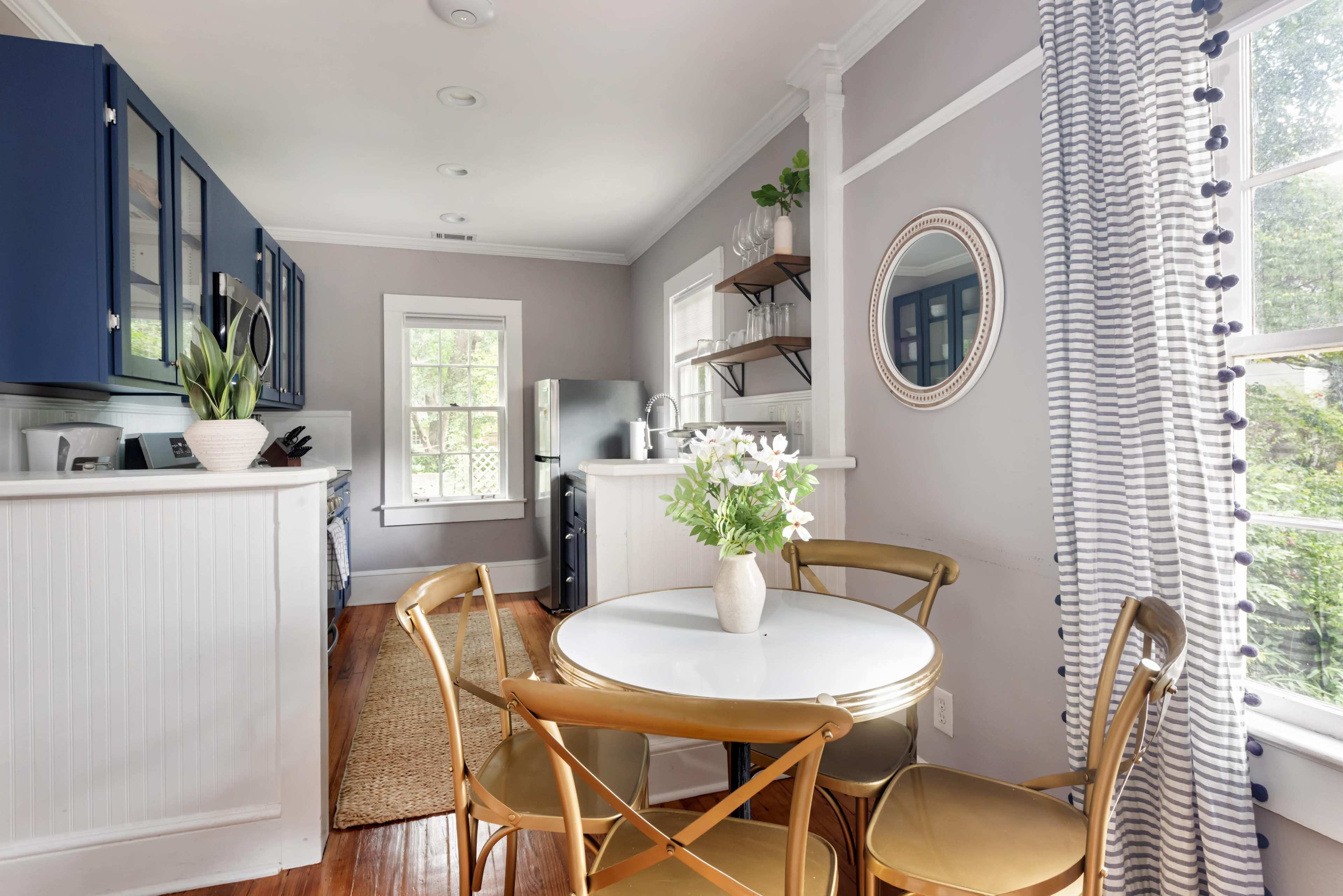 The image shows a modern kitchen with blue cabinetry, a circular white dining table surrounded by gold chairs, and large windows allowing natural light to enter.