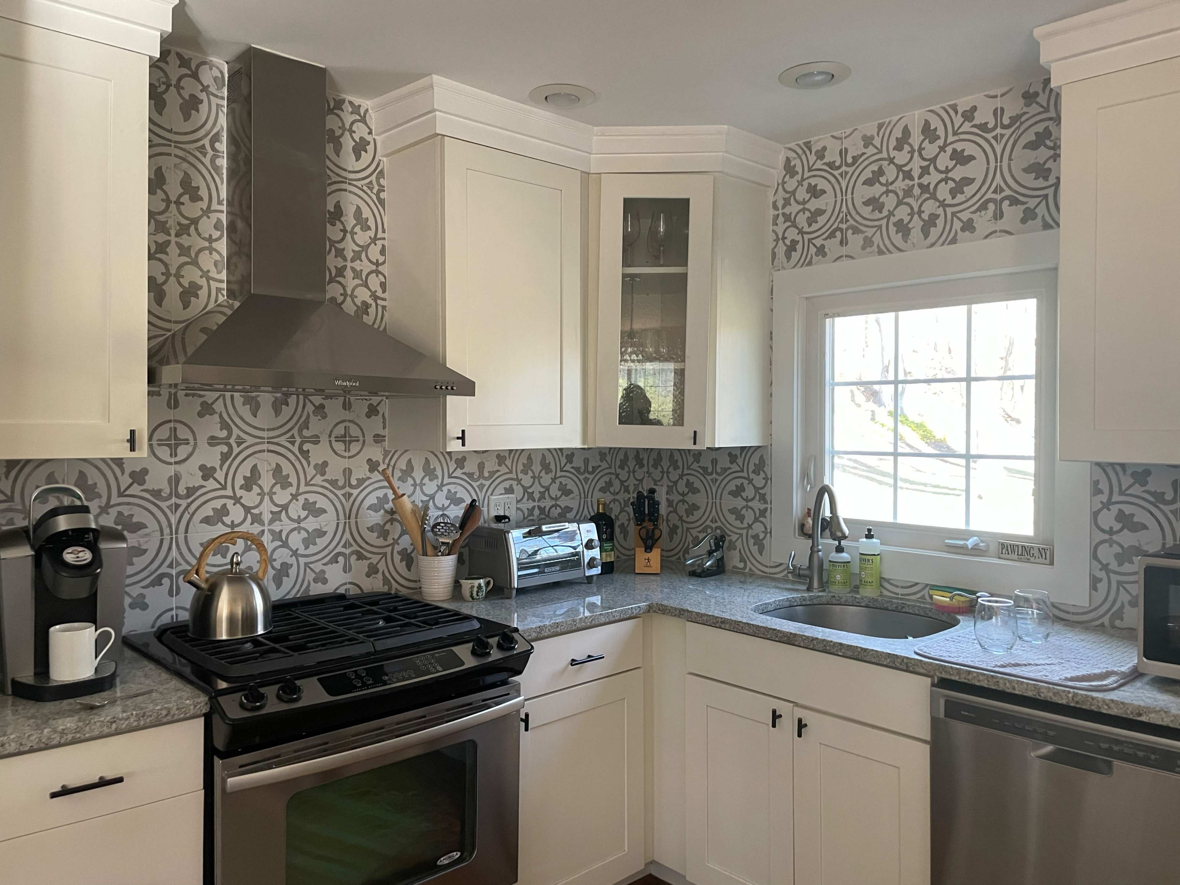 The image shows a modern kitchen featuring white cabinets, patterned wallpaper, a stainless steel stove, a sink, and a coffee maker on the counter.