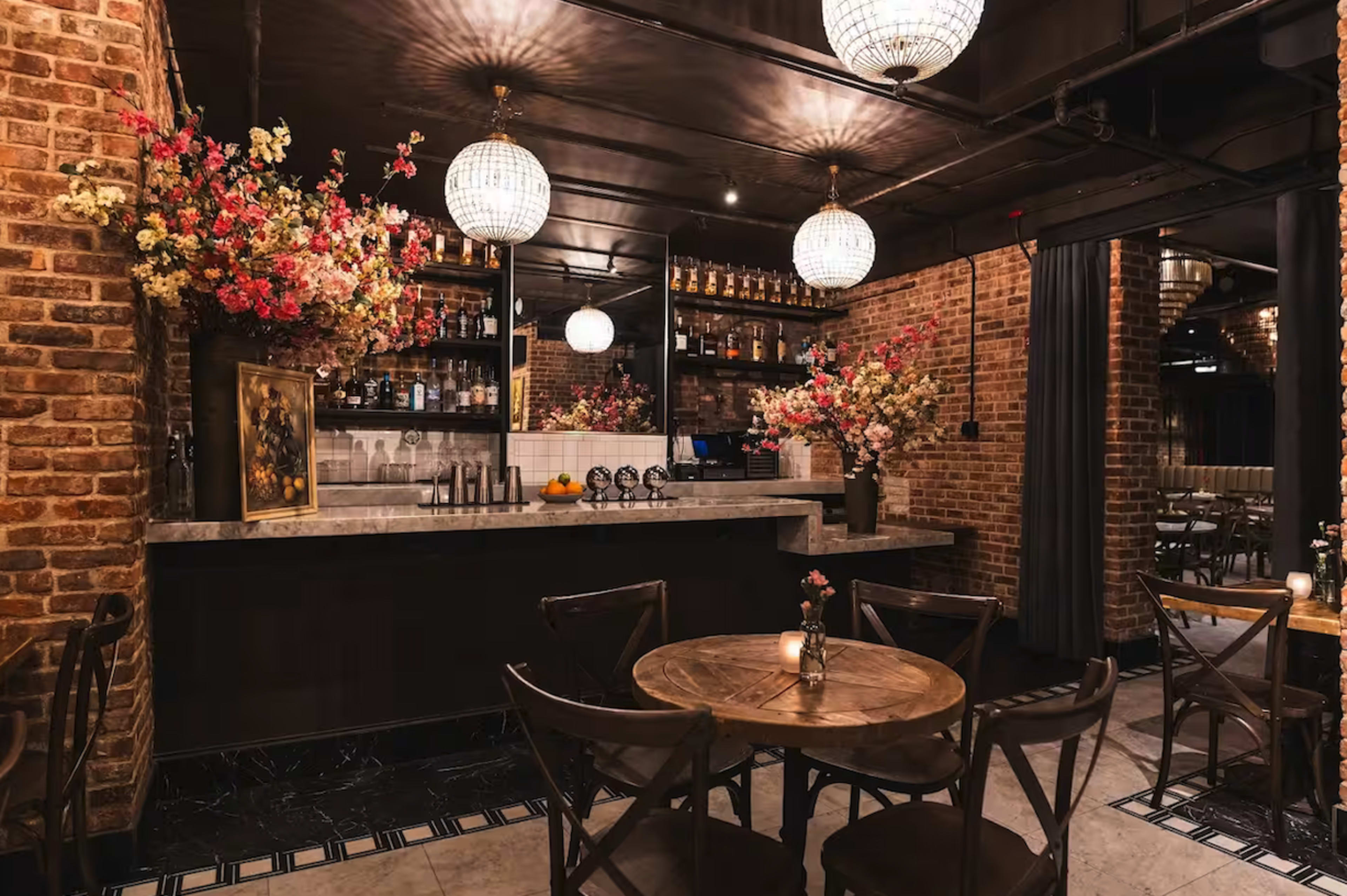 The image shows a cozy restaurant interior featuring a bar with a marble countertop, wooden tables and chairs, and decorative flower arrangements in a brick-walled setting.