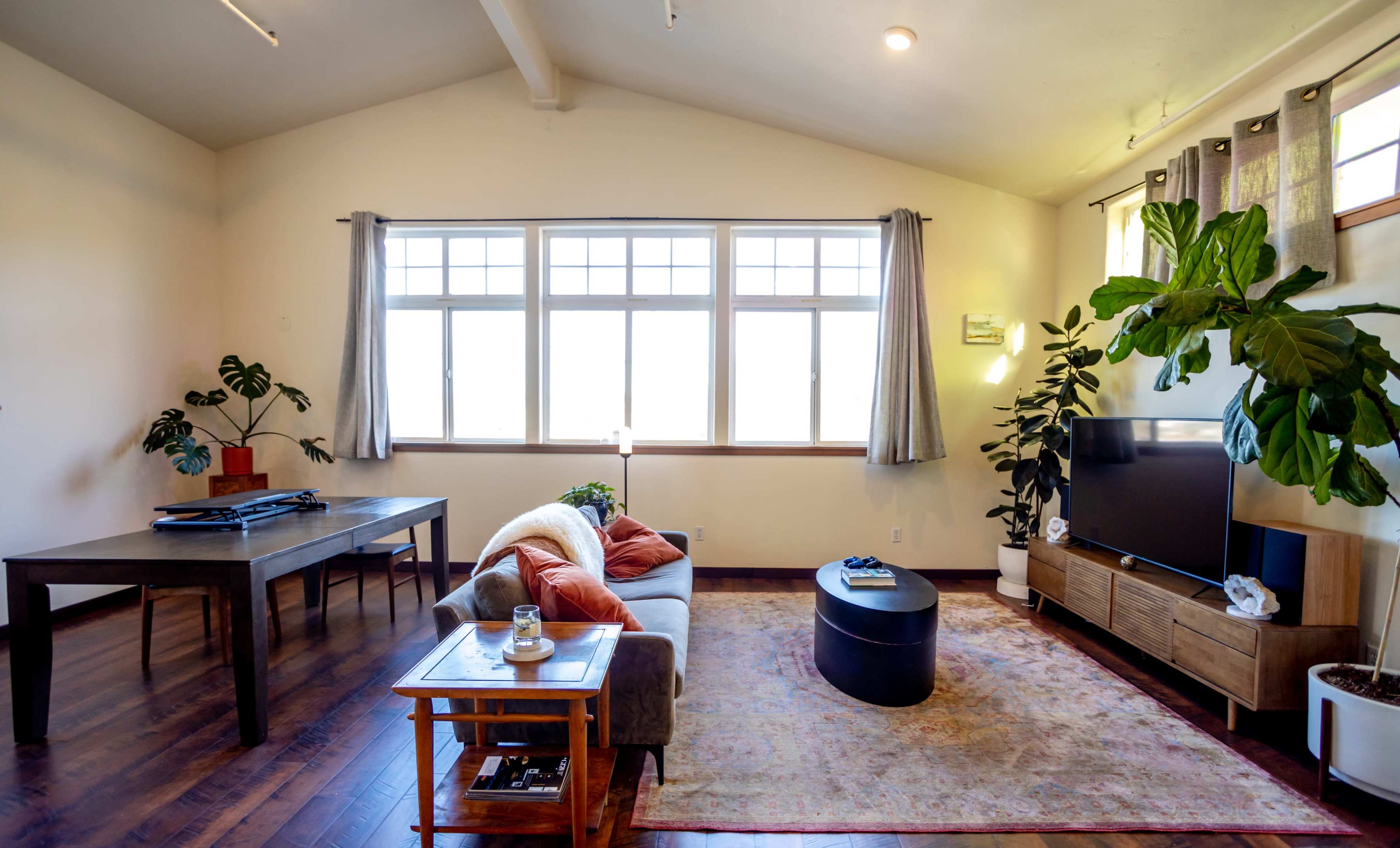 A cozy living room features a sofa with orange and gray cushions, a wooden coffee table, and a television on a console under large windows with sheer curtains.