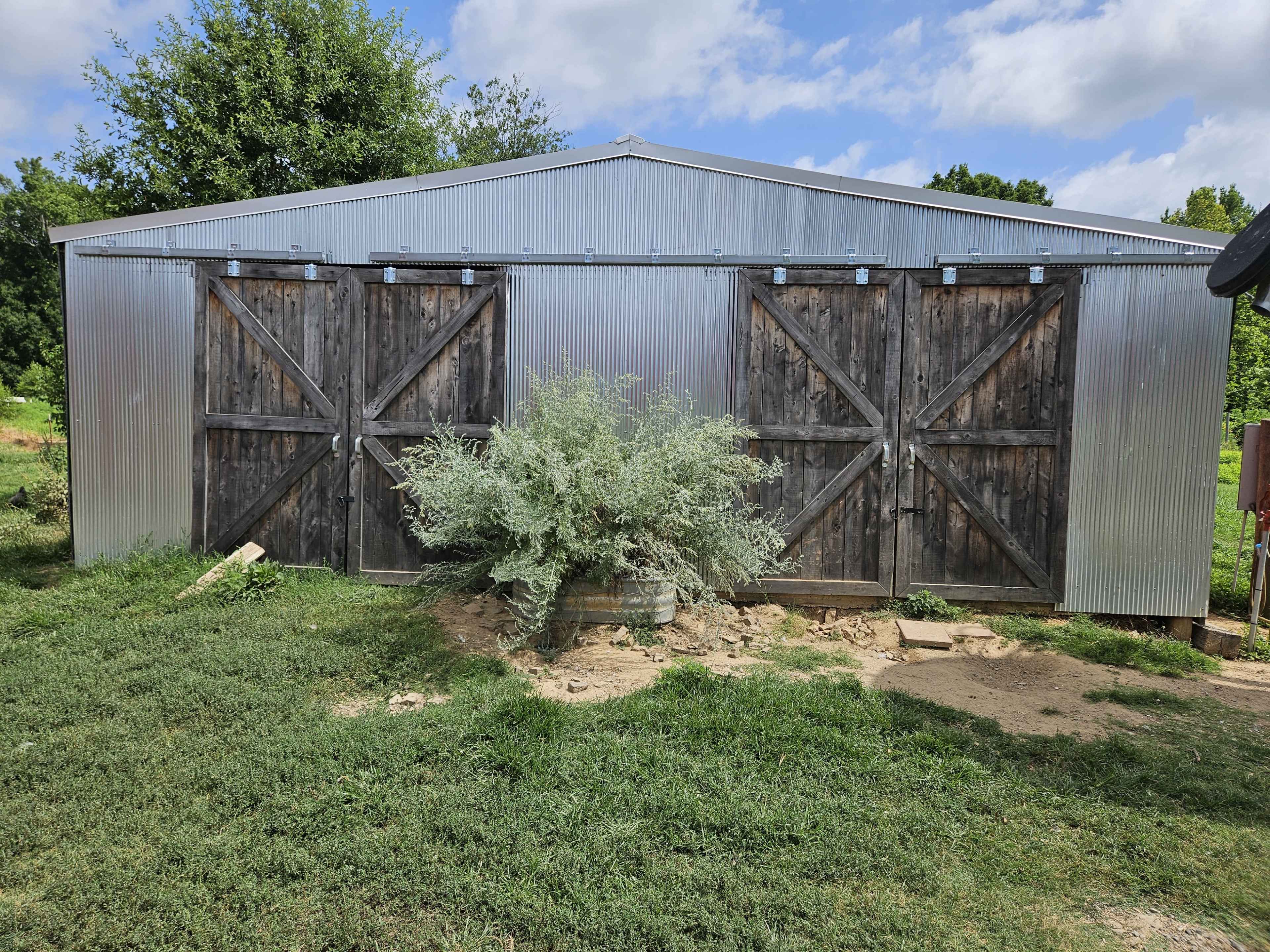 The image shows a large metal barn with wooden double doors and a bush in front, set against a grassy area under a partly cloudy sky.