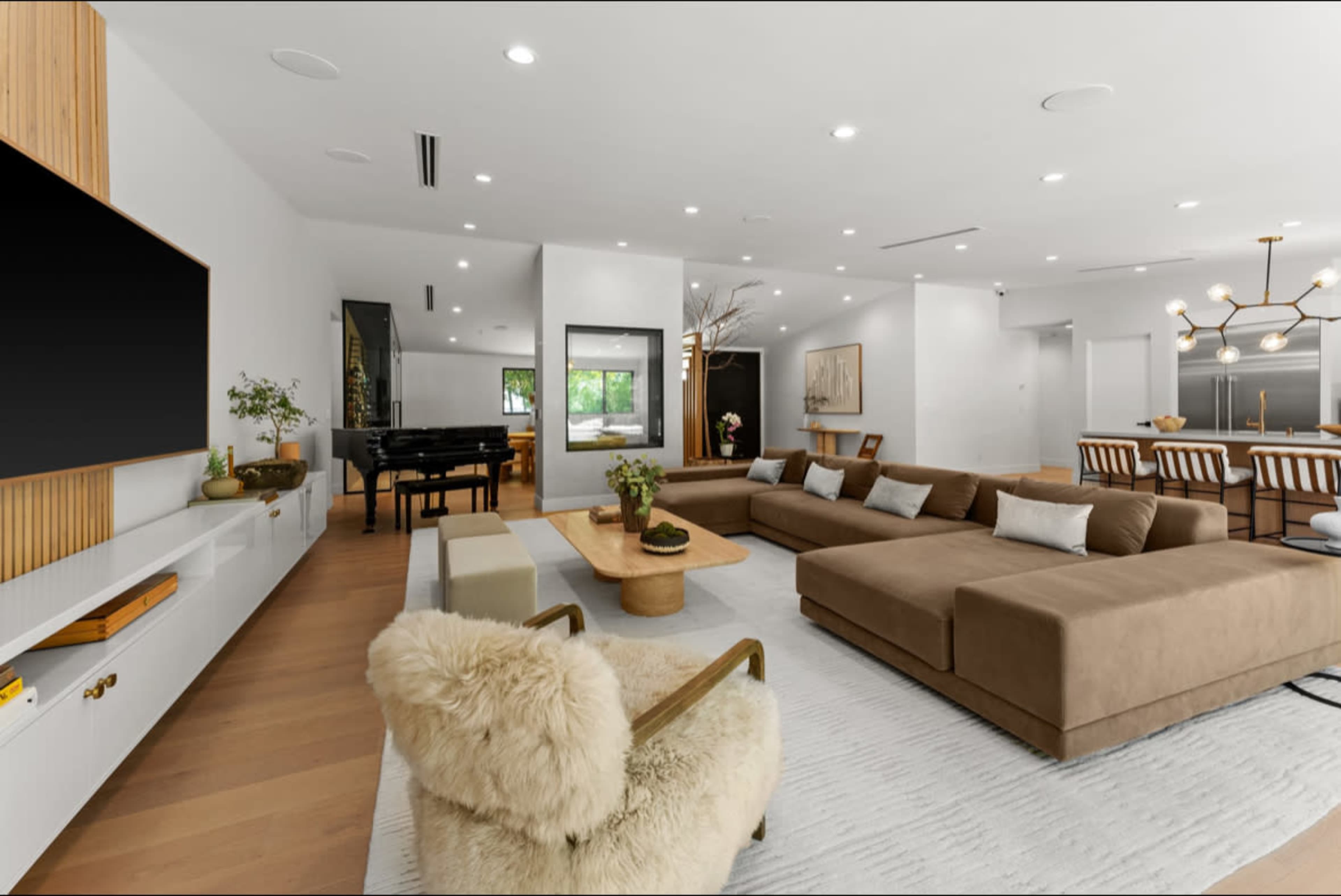 A modern living room featuring a large brown sectional sofa, a wooden coffee table, a plush armchair, and a piano in the background, with ample lighting and minimalist decor.