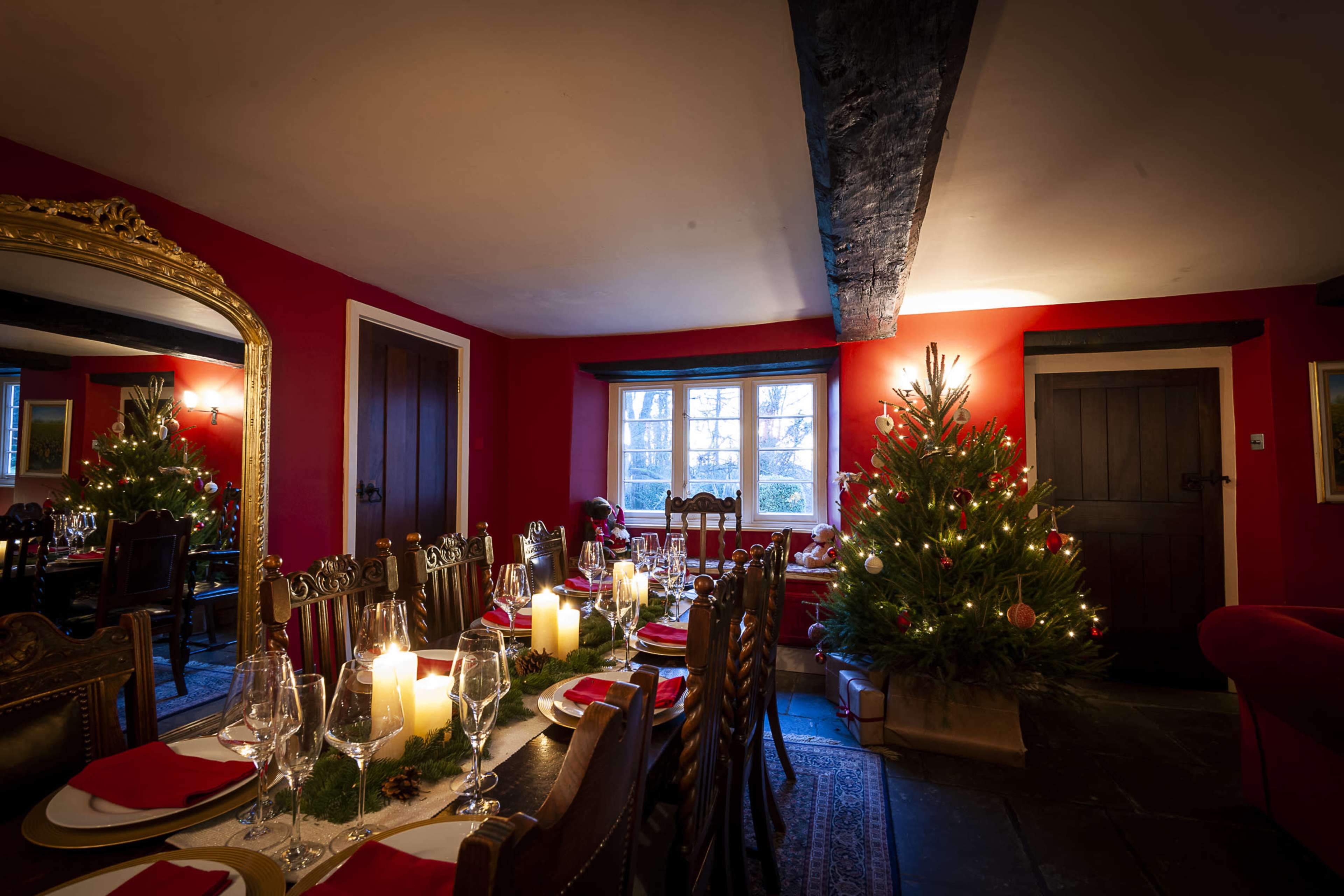 A festively decorated dining room features a long table set for a meal, with candles and a Christmas tree illuminated in the corner.