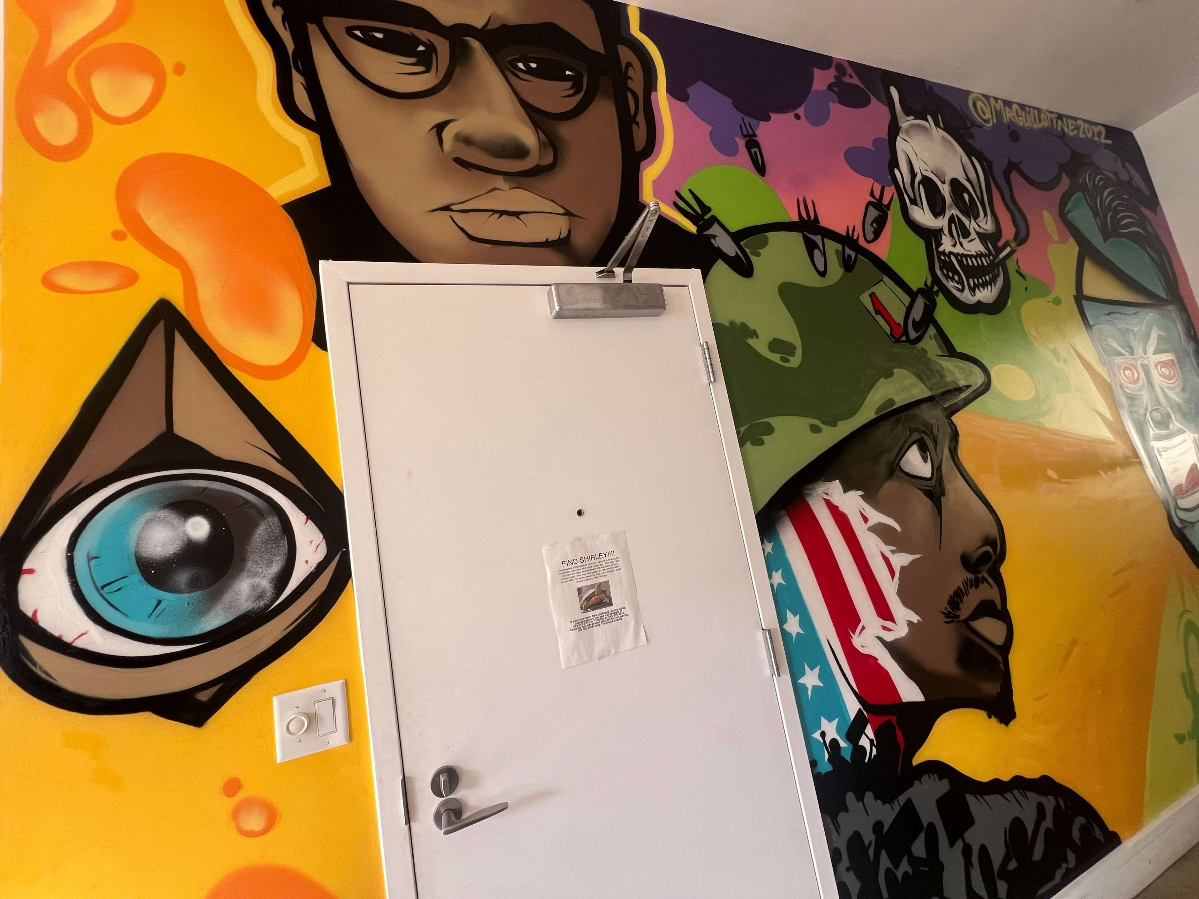 A brightly colored mural on a wall featuring various graphic elements, including a stylized eye, a figure wearing a military helmet, and a face adorned with an American flag.