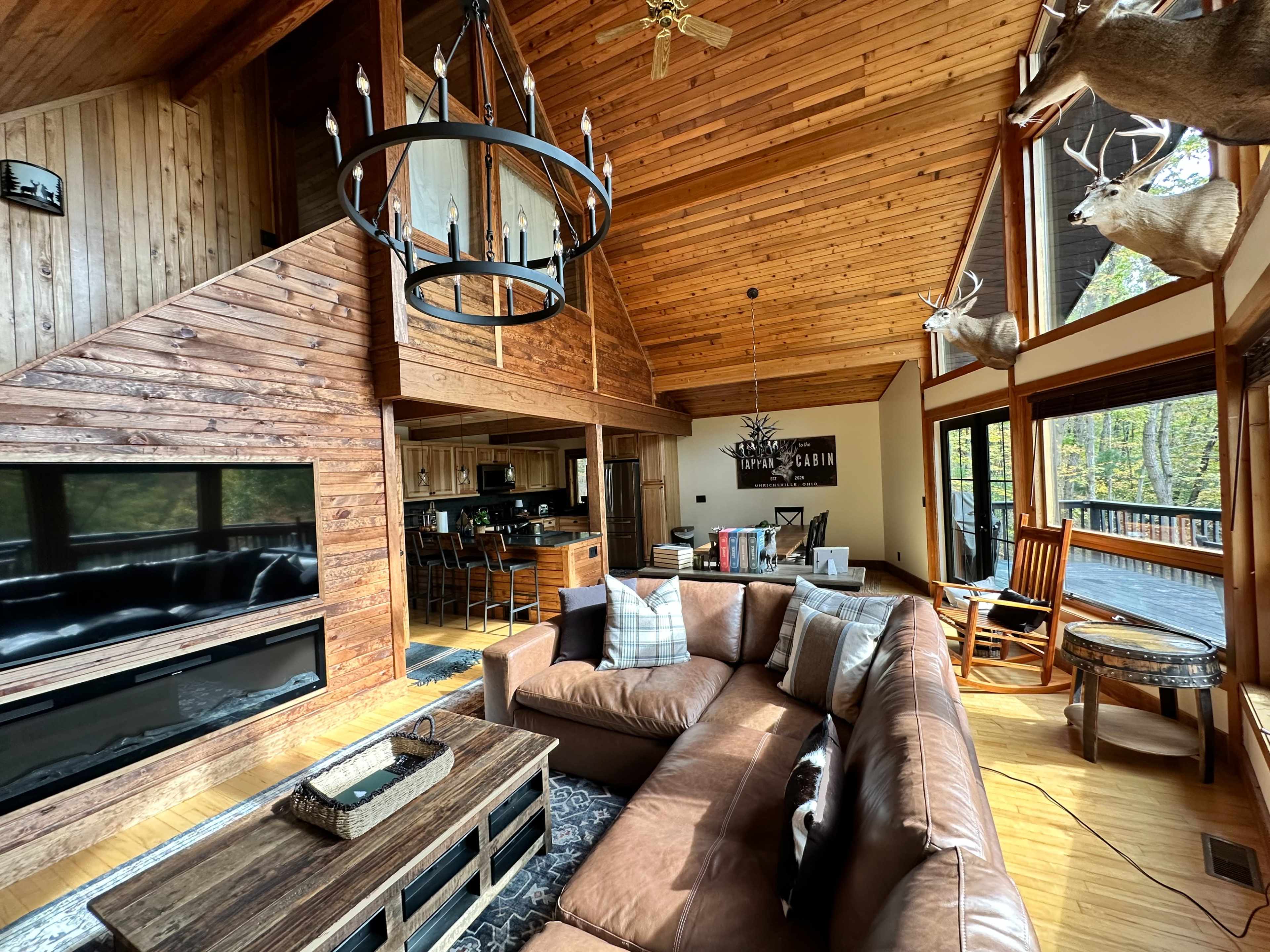 The image shows a spacious living room with wooden walls and ceilings, featuring a large leather sectional sofa, a flat-screen TV, and decorative deer heads mounted on the walls.