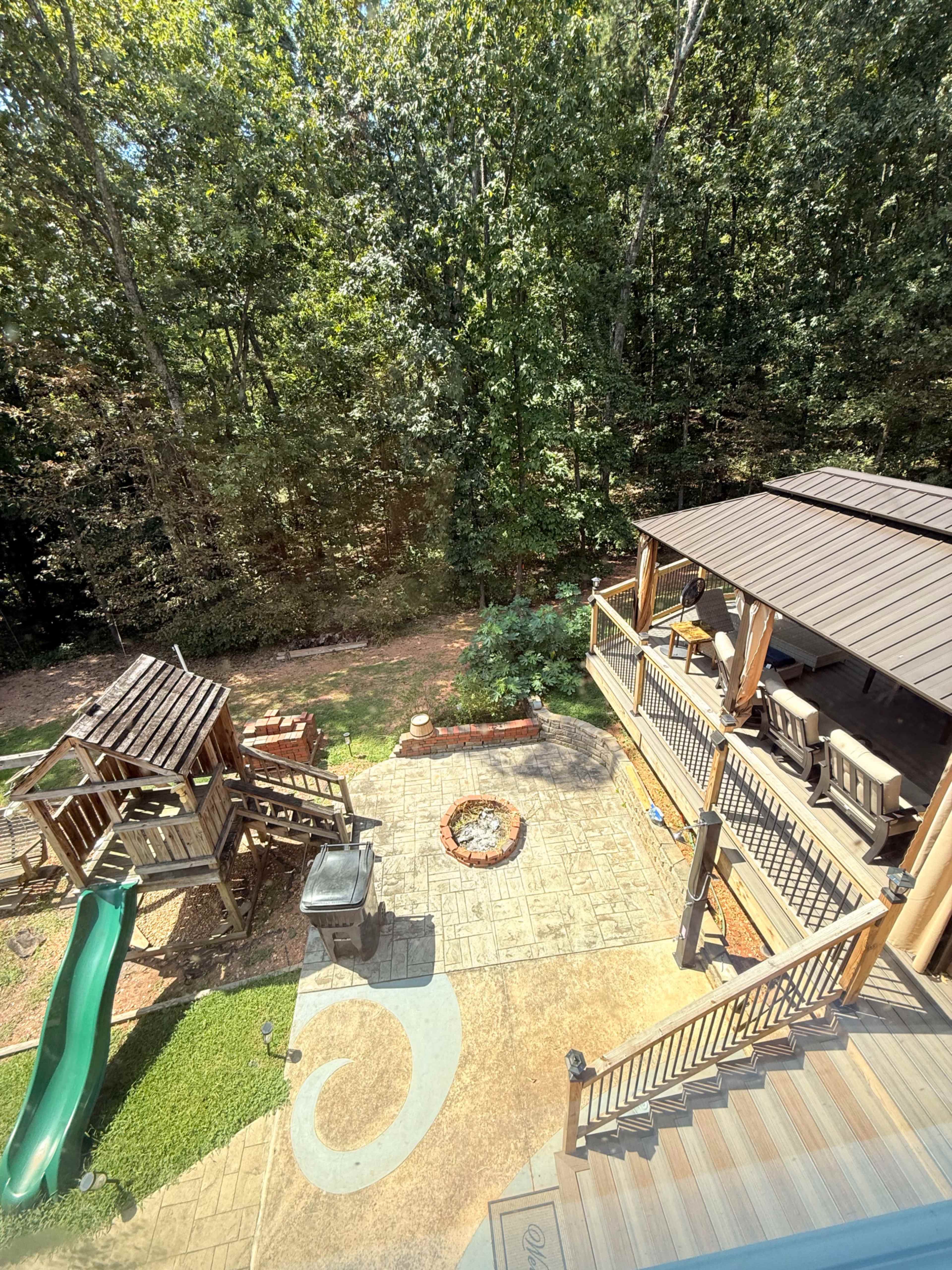 The image shows a backyard scene with a patio area, a fire pit, a children's playset with a slide, and a dense tree line in the background.