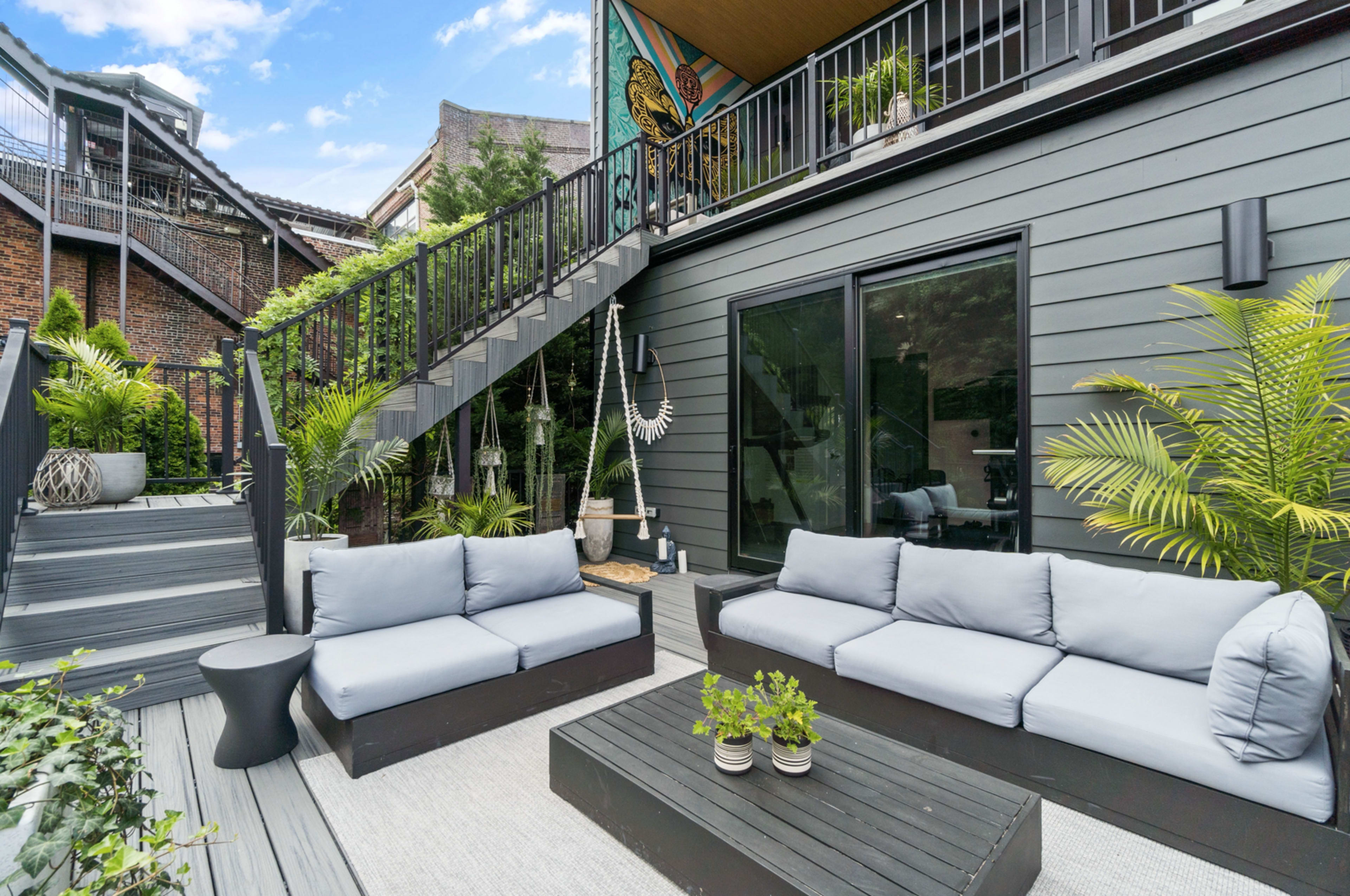 A modern outdoor seating area features gray sofas and a coffee table amidst greenery and stairs leading to a second level.