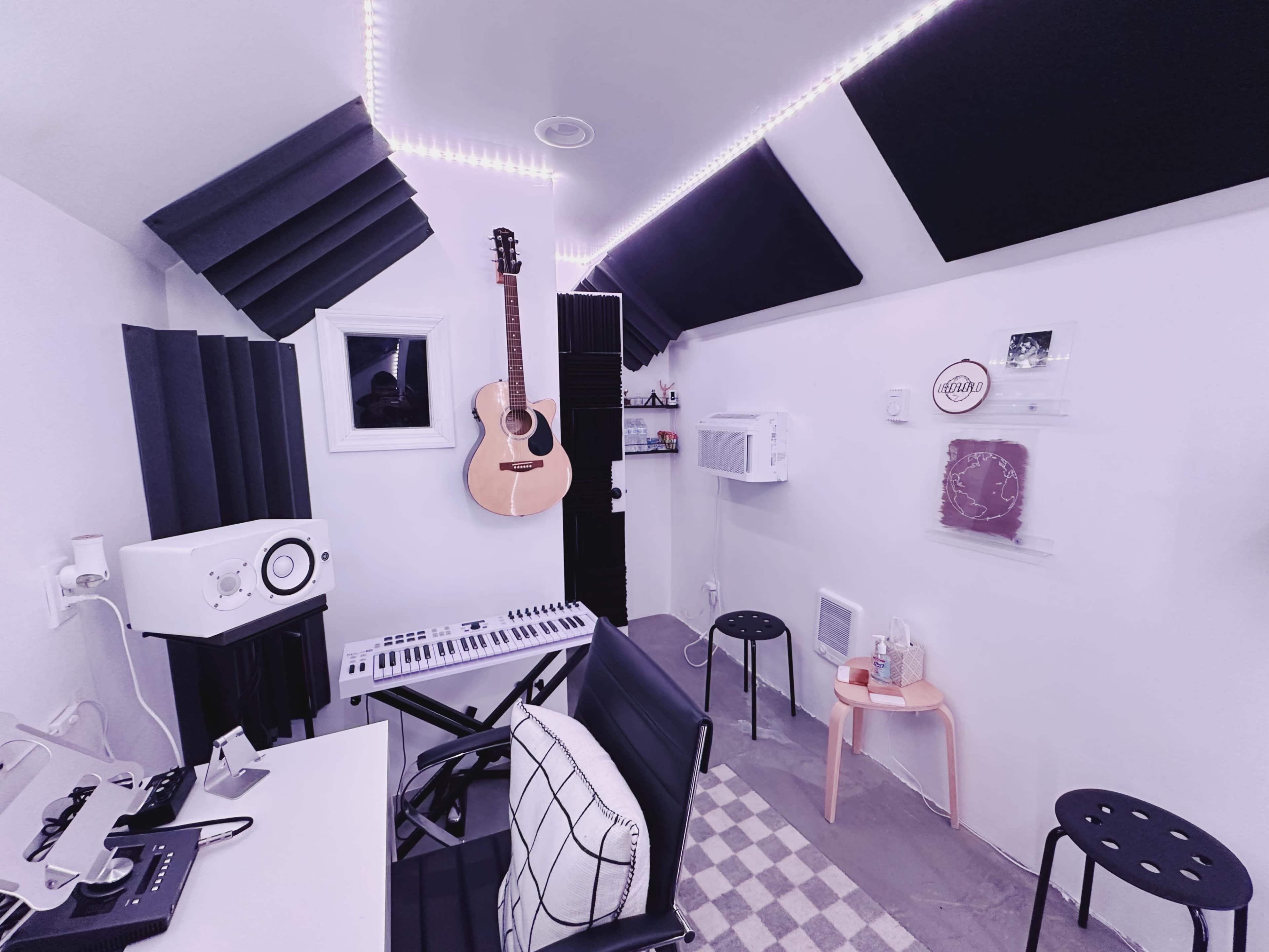 A small music studio with a guitar hanging on the wall, a keyboard on a desk, and acoustic panels on the ceiling.