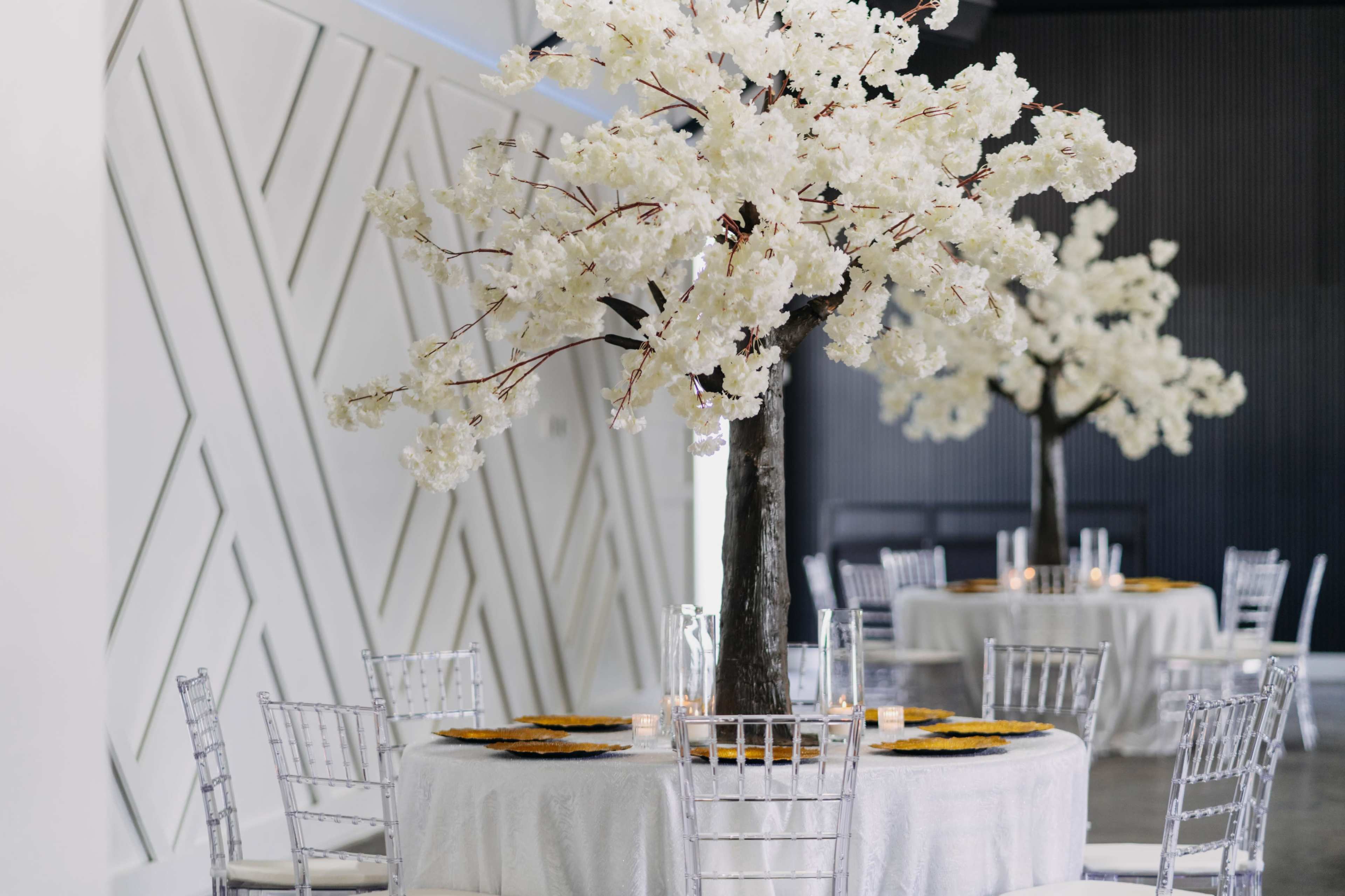 The image shows an elegant event space featuring round tables with white tablecloths, clear chiavari chairs, and tall floral arrangements of white blossoms at the center.