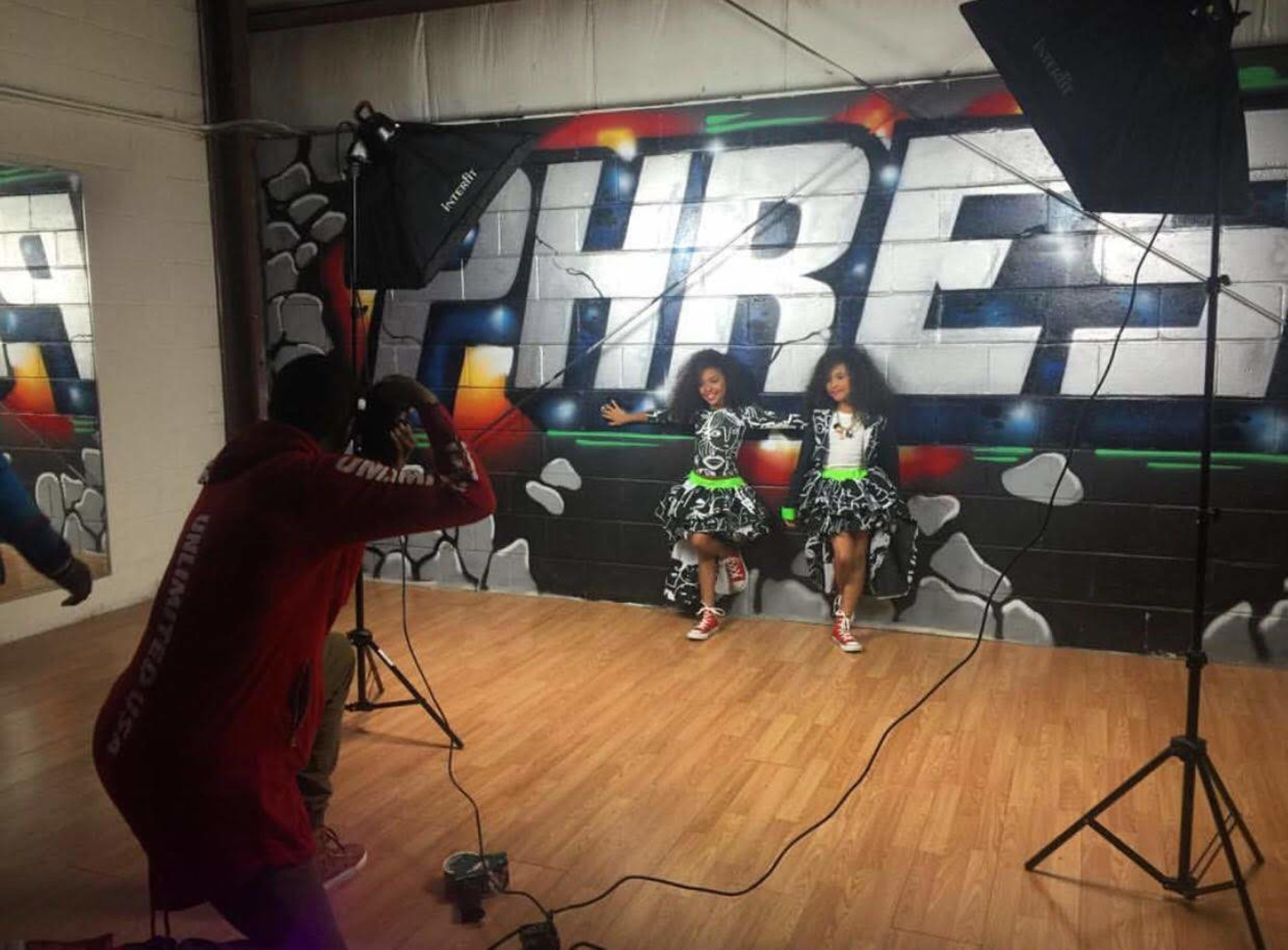 Two dancers pose in colorful outfits against a graffiti-covered wall while a photographer captures the moment in a dance studio.