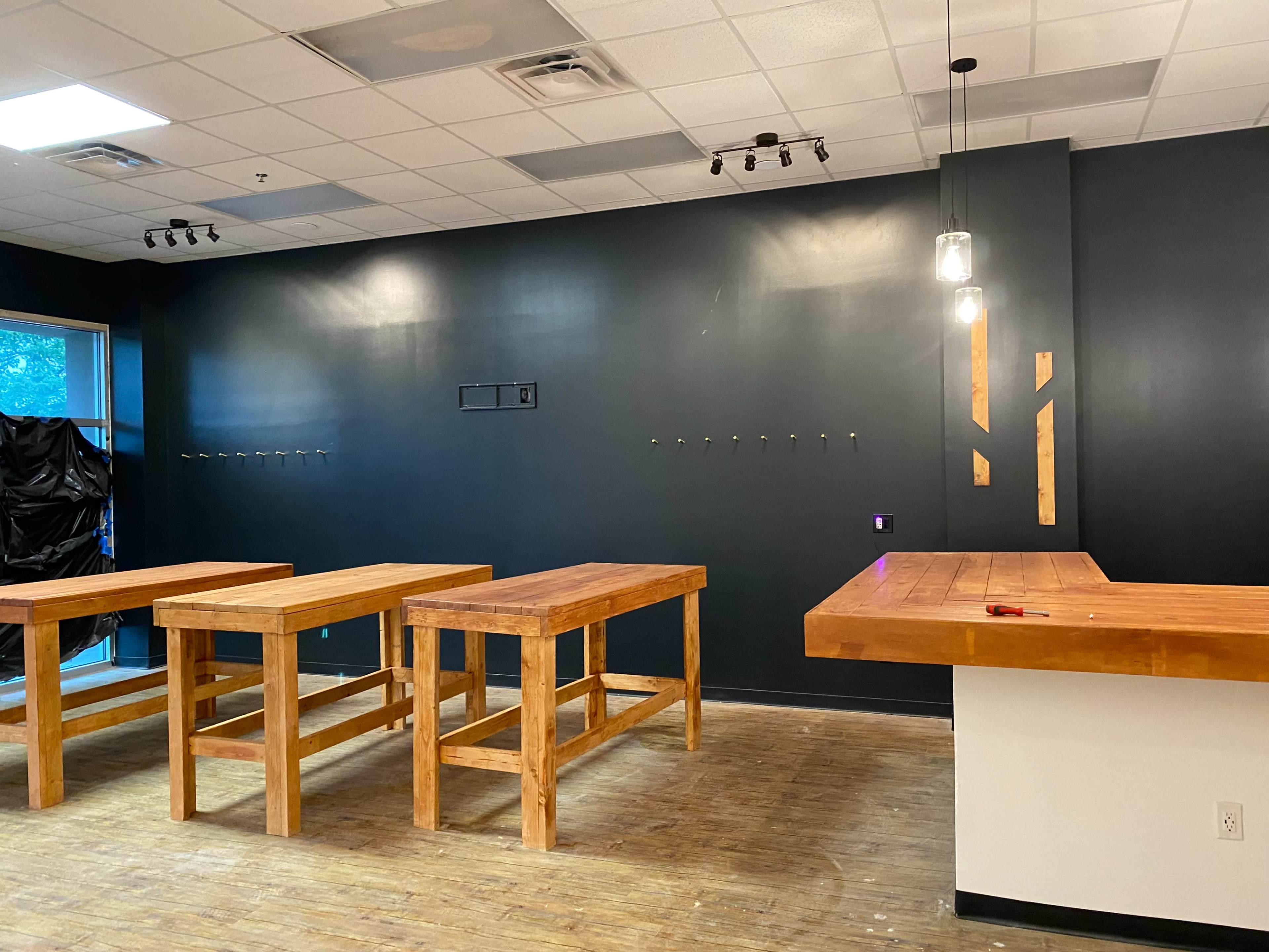 An interior space with several wooden tables arranged in a row and a dark wall with hooks, illuminated by overhead lighting.