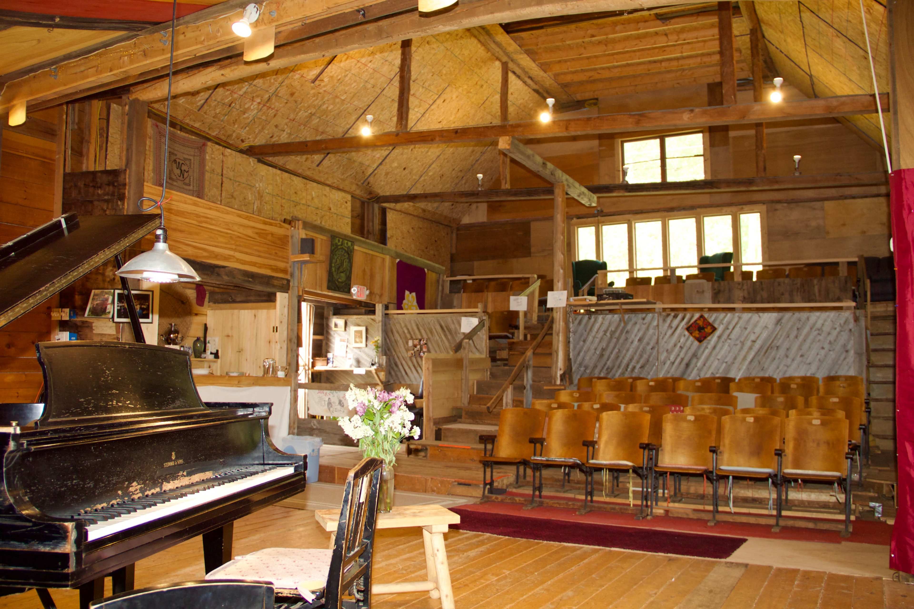 The image shows a wooden interior of a small performance space featuring a grand piano in the foreground and rows of wooden chairs arranged facing a stage in the background.