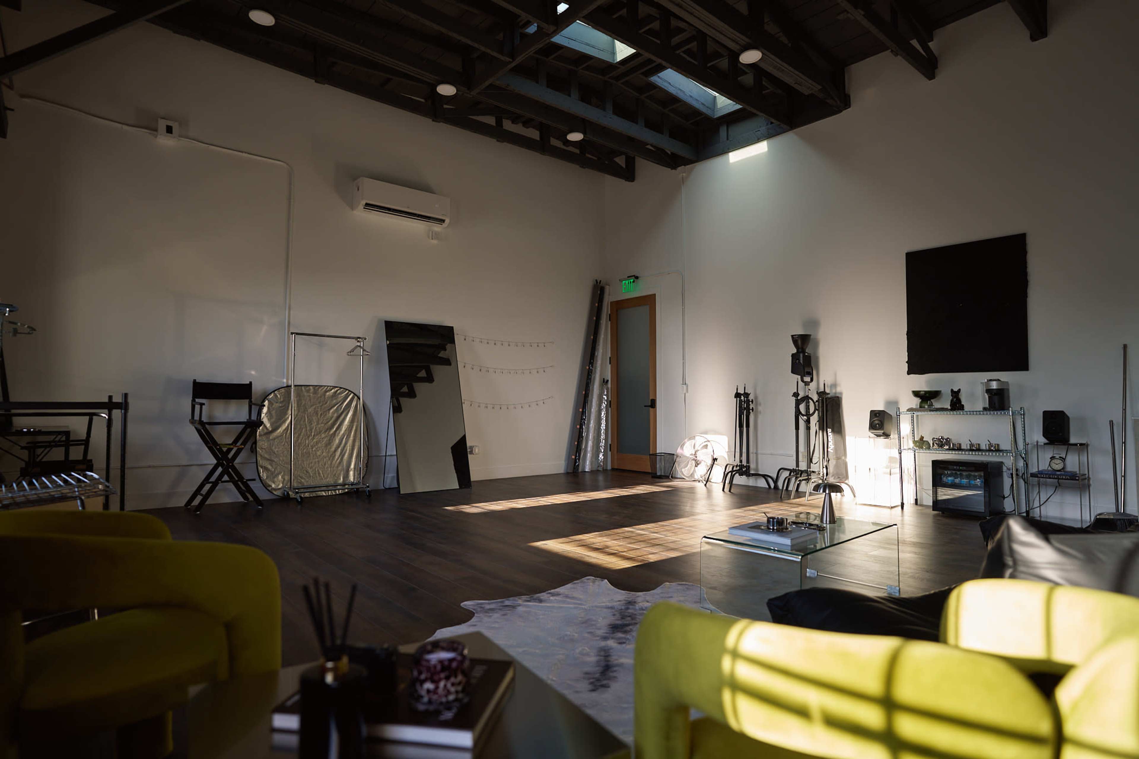 The image depicts a spacious, well-lit interior room with wooden flooring, featuring minimal furnishings such as two yellow chairs, a large mirror, and a television on the wall.