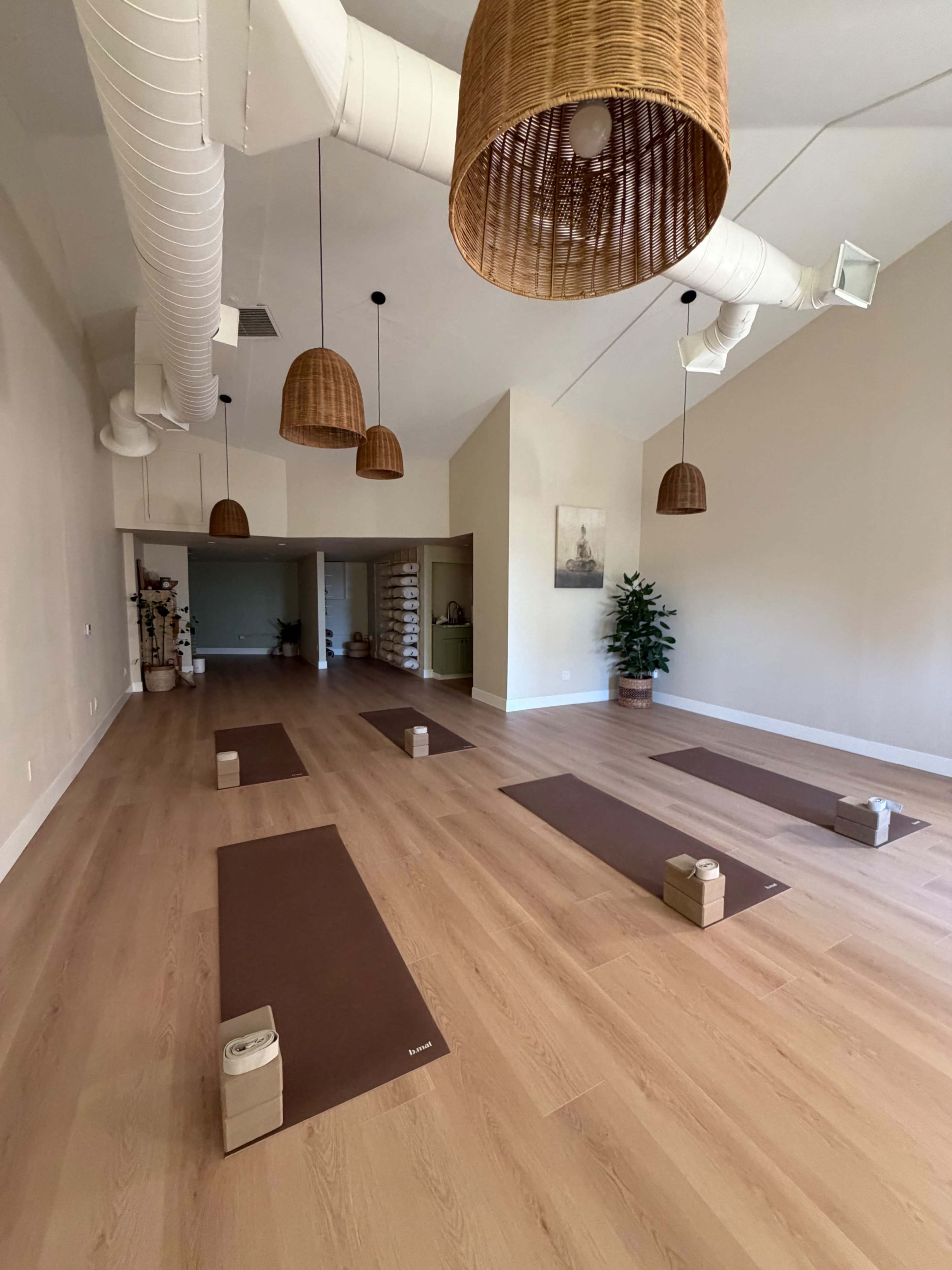 The image shows a spacious yoga studio with wooden floors, several yoga mats arranged on the ground, and hanging wicker light fixtures.