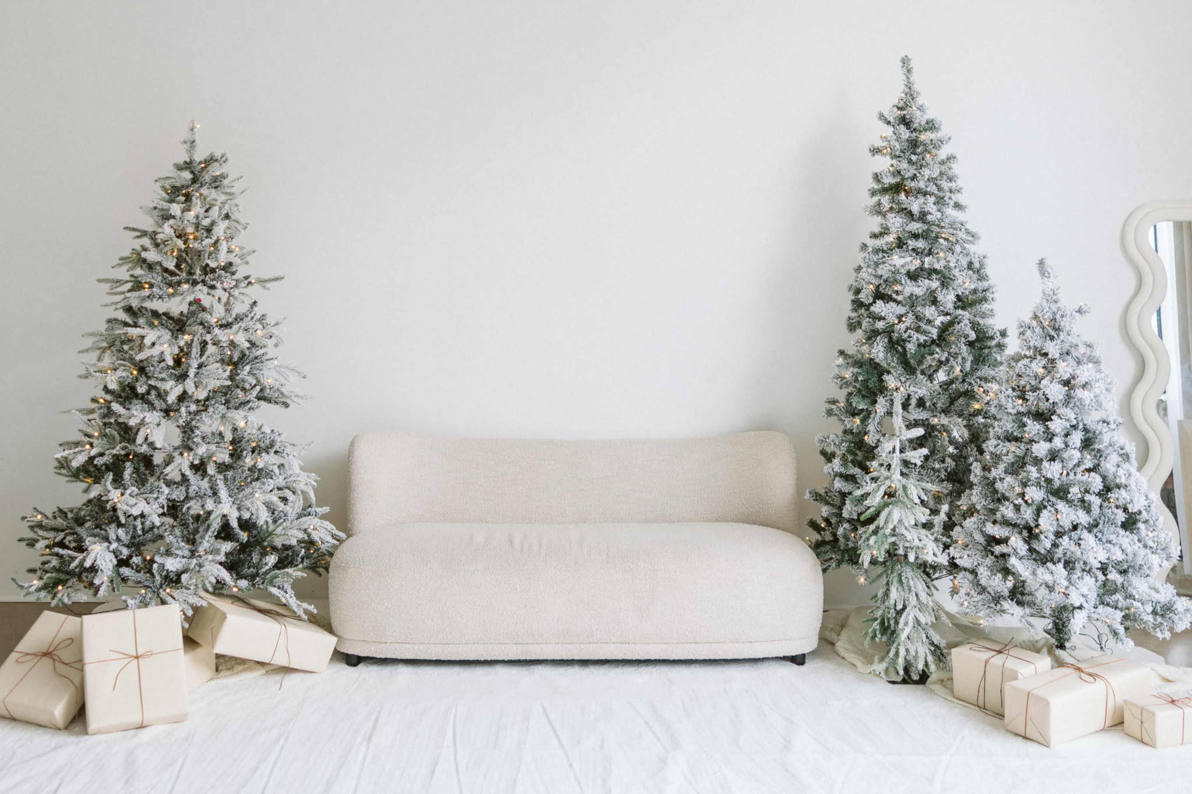 The image shows a cozy living space featuring a light-colored couch flanked by two frosted Christmas trees and neatly wrapped gifts on a white floor.