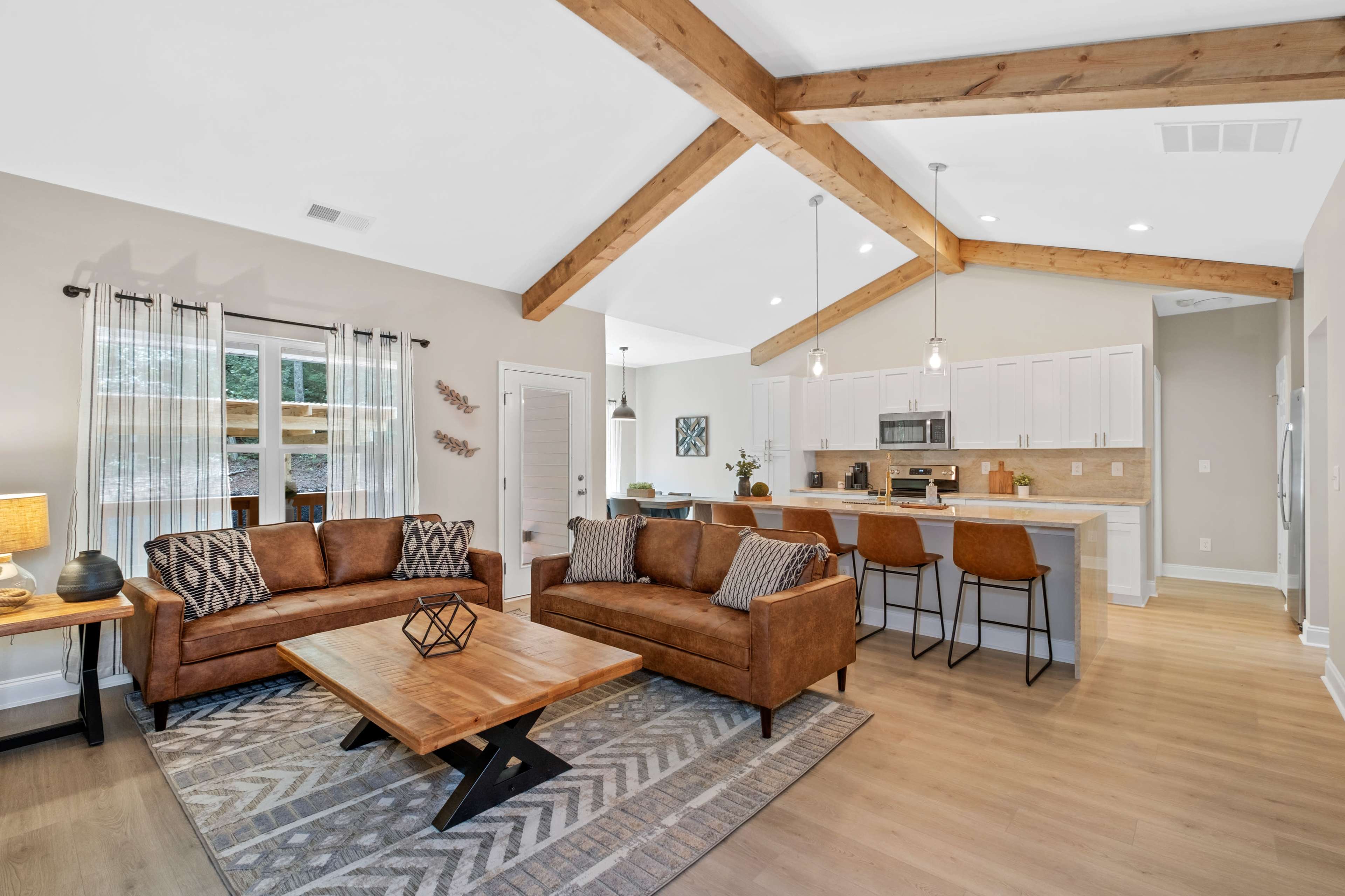 A modern living room with two brown sofas, a wooden coffee table, and a kitchen area in the background featuring white cabinets and stainless steel appliances.