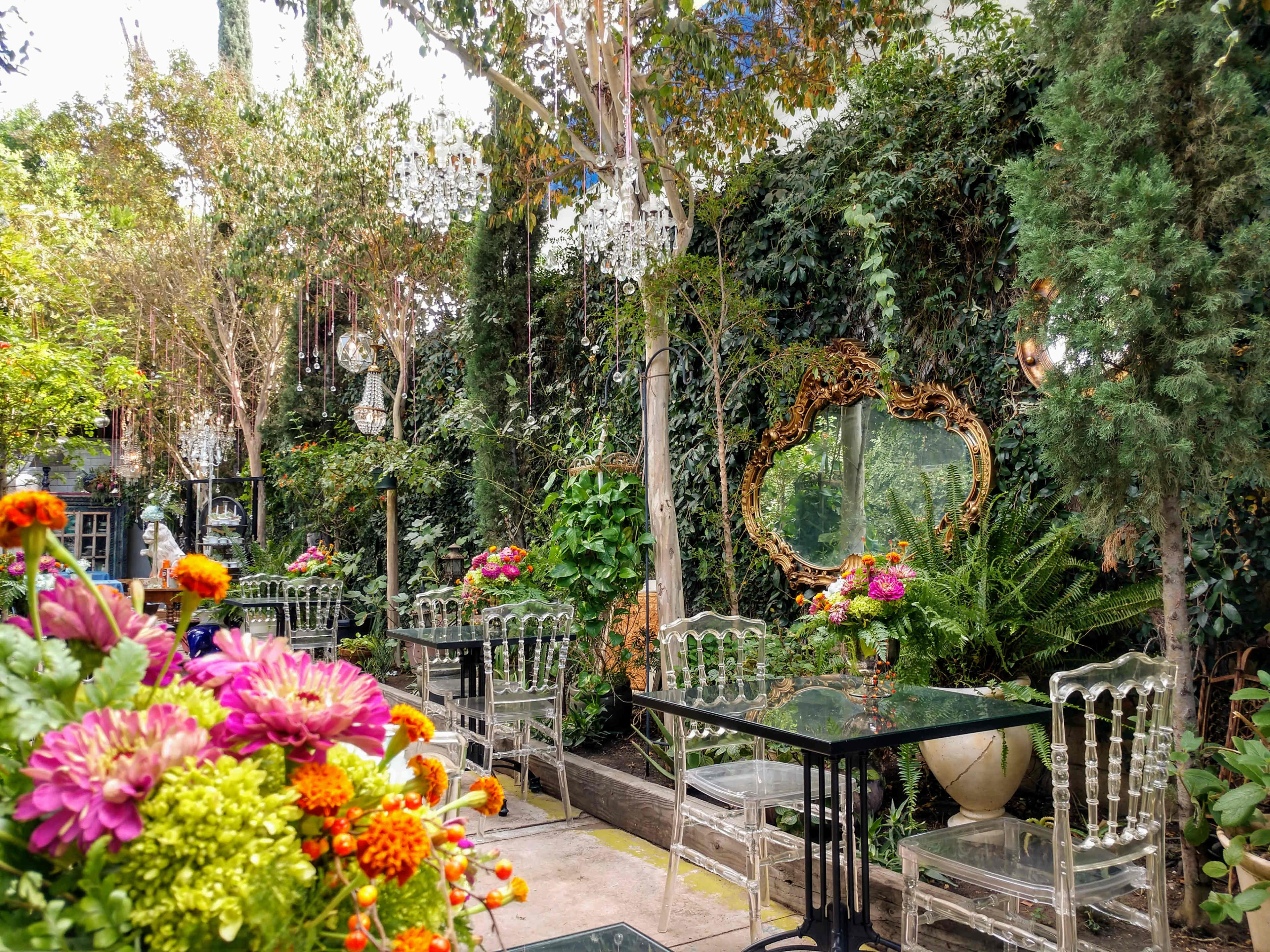 The image shows a lush garden patio with transparent acrylic chairs arranged around black tables, surrounded by vibrant flowers and greenery, and featuring a decorative mirror on a wall covered in plants.