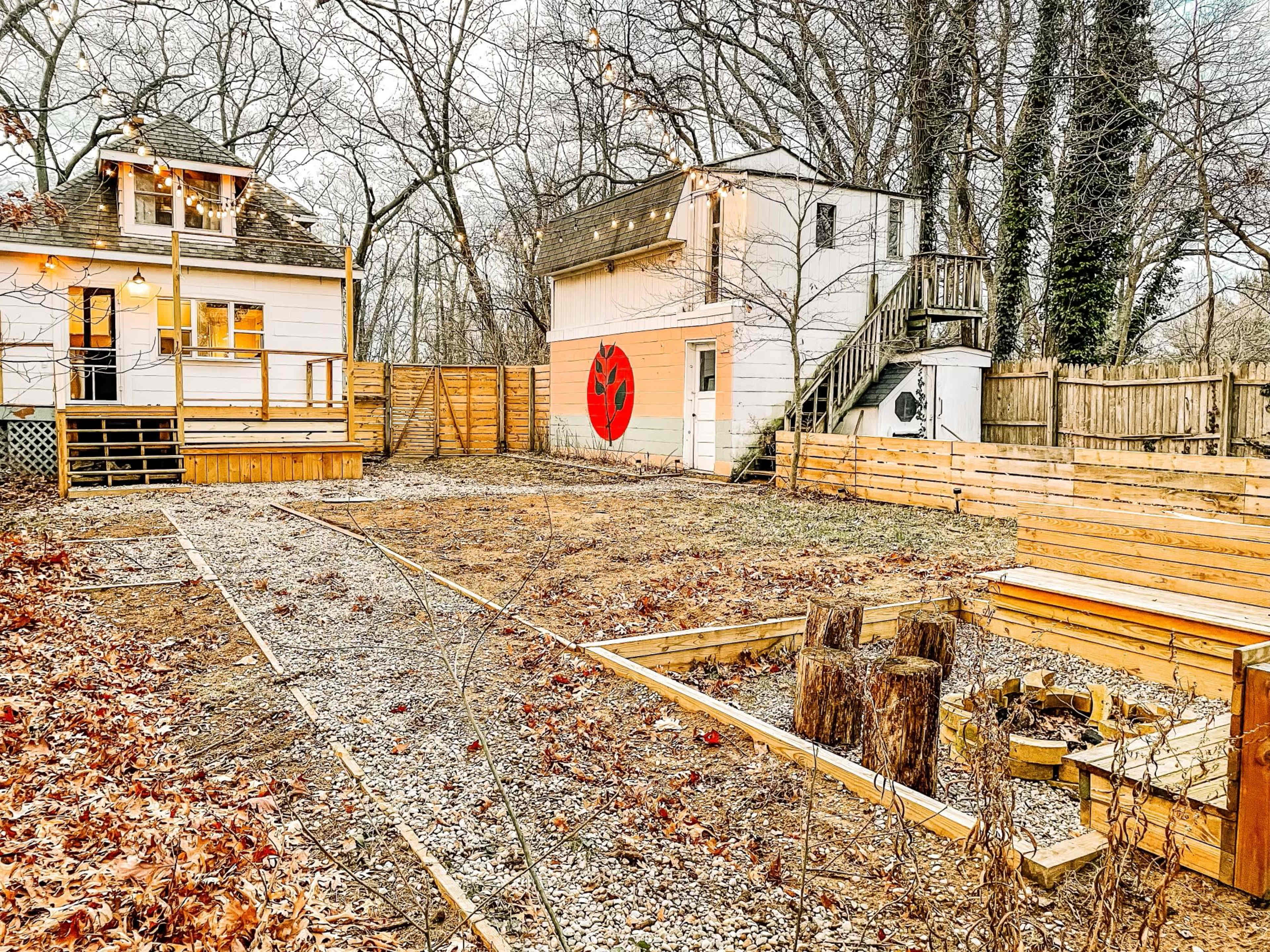 A vacant backyard with gravel and wooden borders, featuring two buildings, one with a red mural, and a set of logs arranged in a circle.
