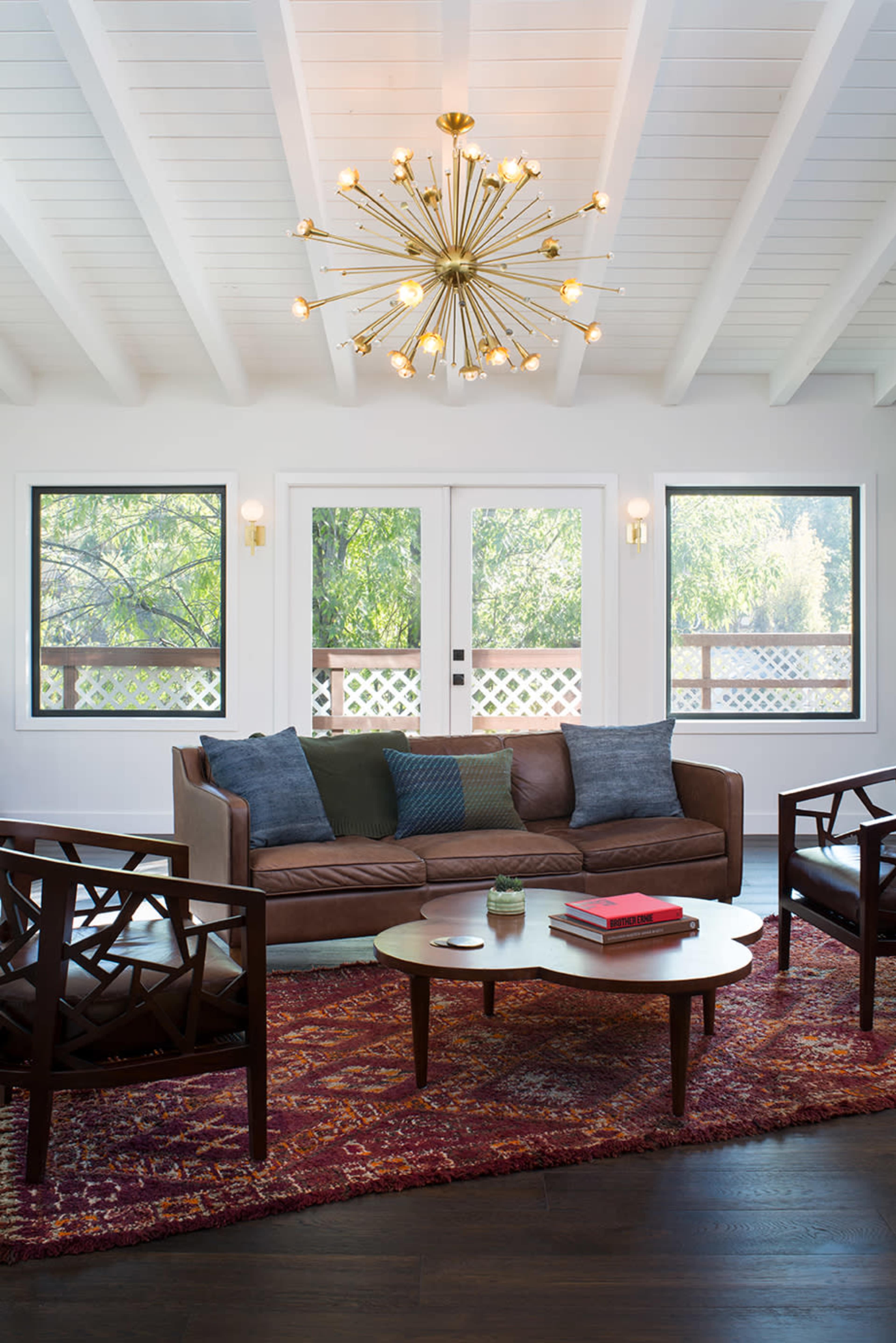 The living room features a brown sofa, two wooden chairs, a round coffee table, and large windows overlooking greenery, all illuminated by a starburst chandelier.