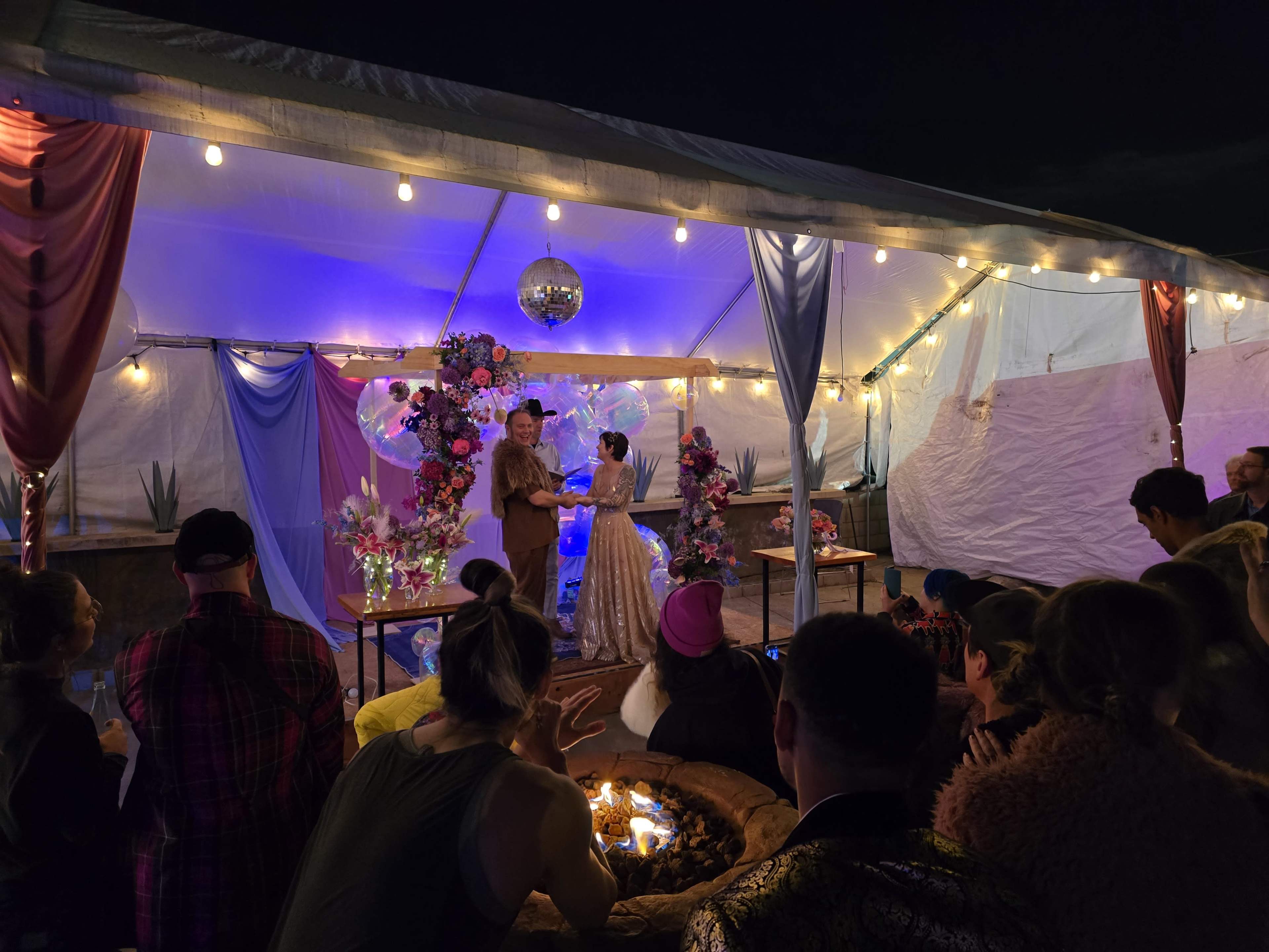 A couple is exchanging vows under a decorated tent with string lights and floral arrangements, while an audience watches.