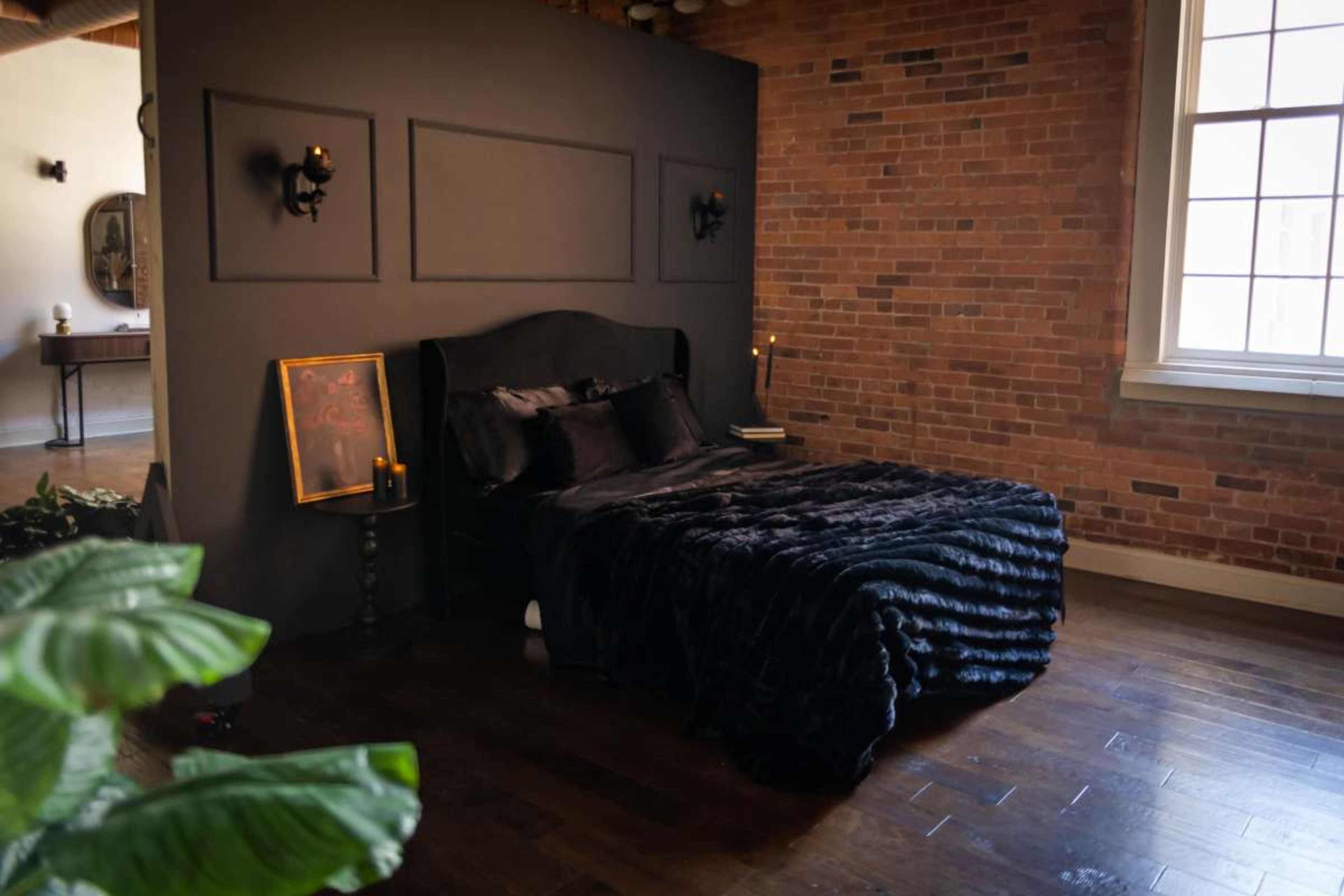 The image shows a bedroom with a darkly painted wall, a black bed adorned with a plush blanket, and exposed brick walls.