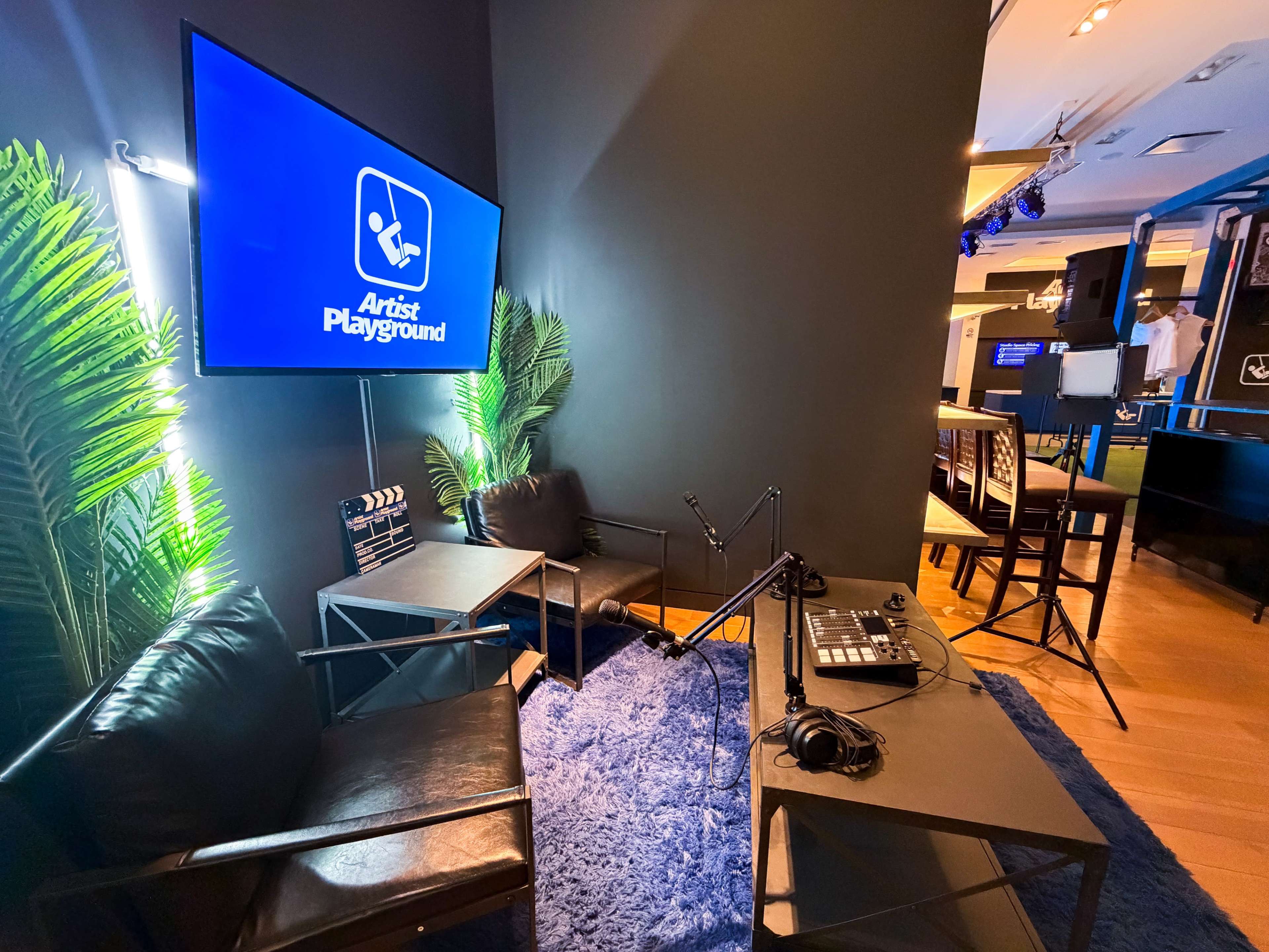 The image shows a small, modern studio setup featuring two black chairs, a microphone on a stand, and a large screen displaying "Artist Playground," surrounded by potted plants and a blue rug.