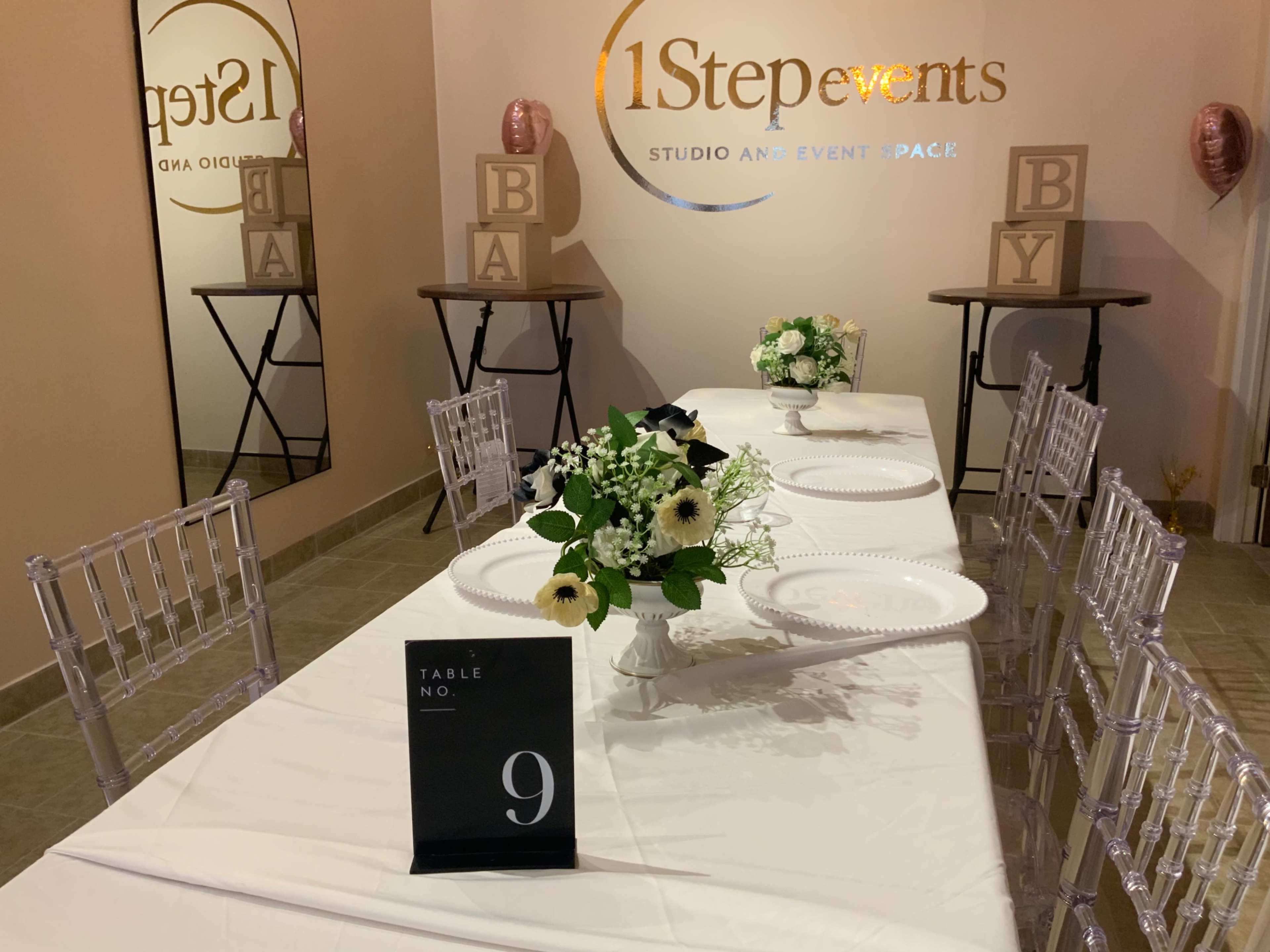 The image shows a decorated event space featuring a long table set with white plates, a floral centerpiece, and a sign indicating "Table No. 9."