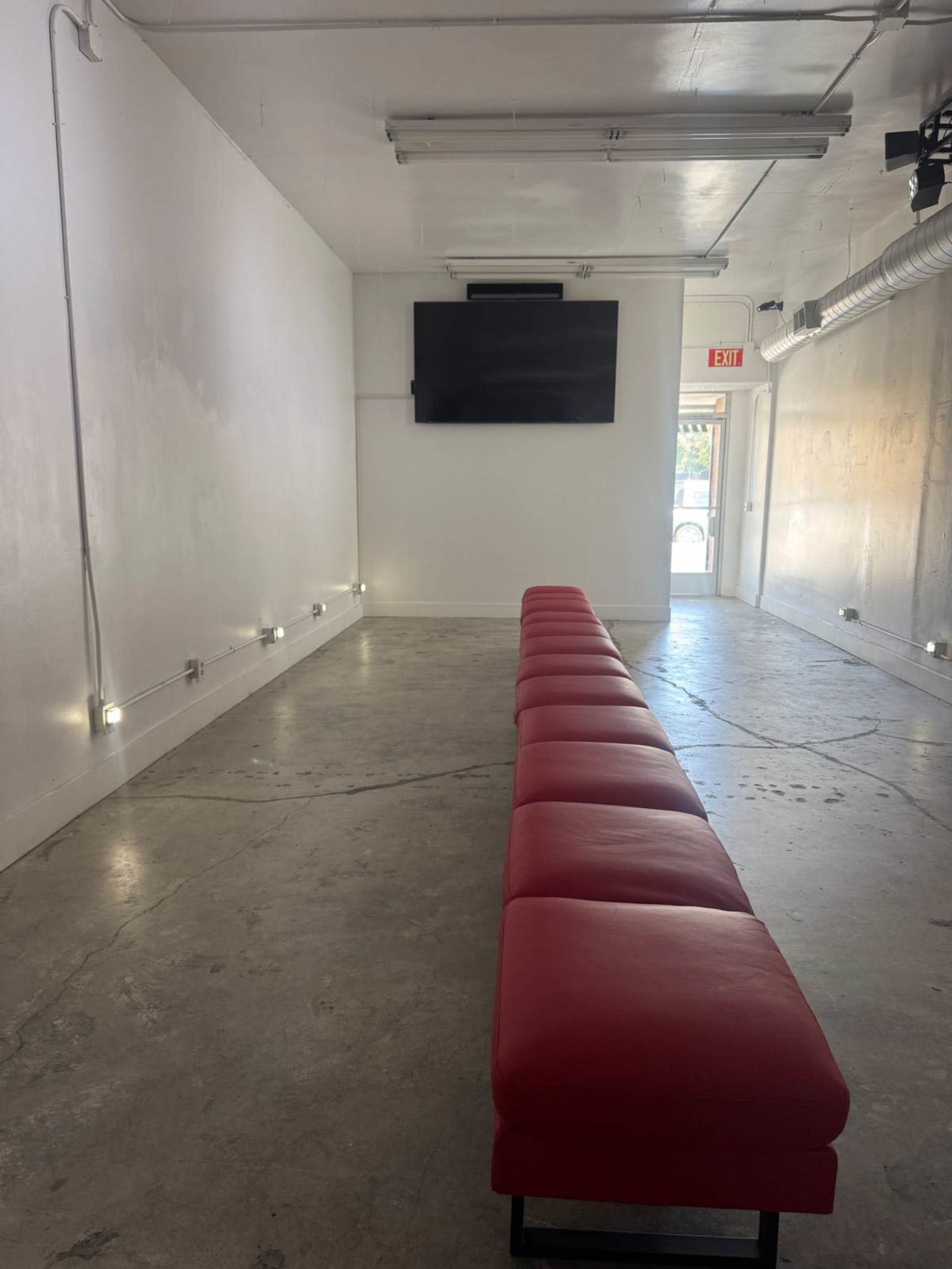 The image shows a minimalist, unfinished room with a long red bench along the center and a wall-mounted television at one end.