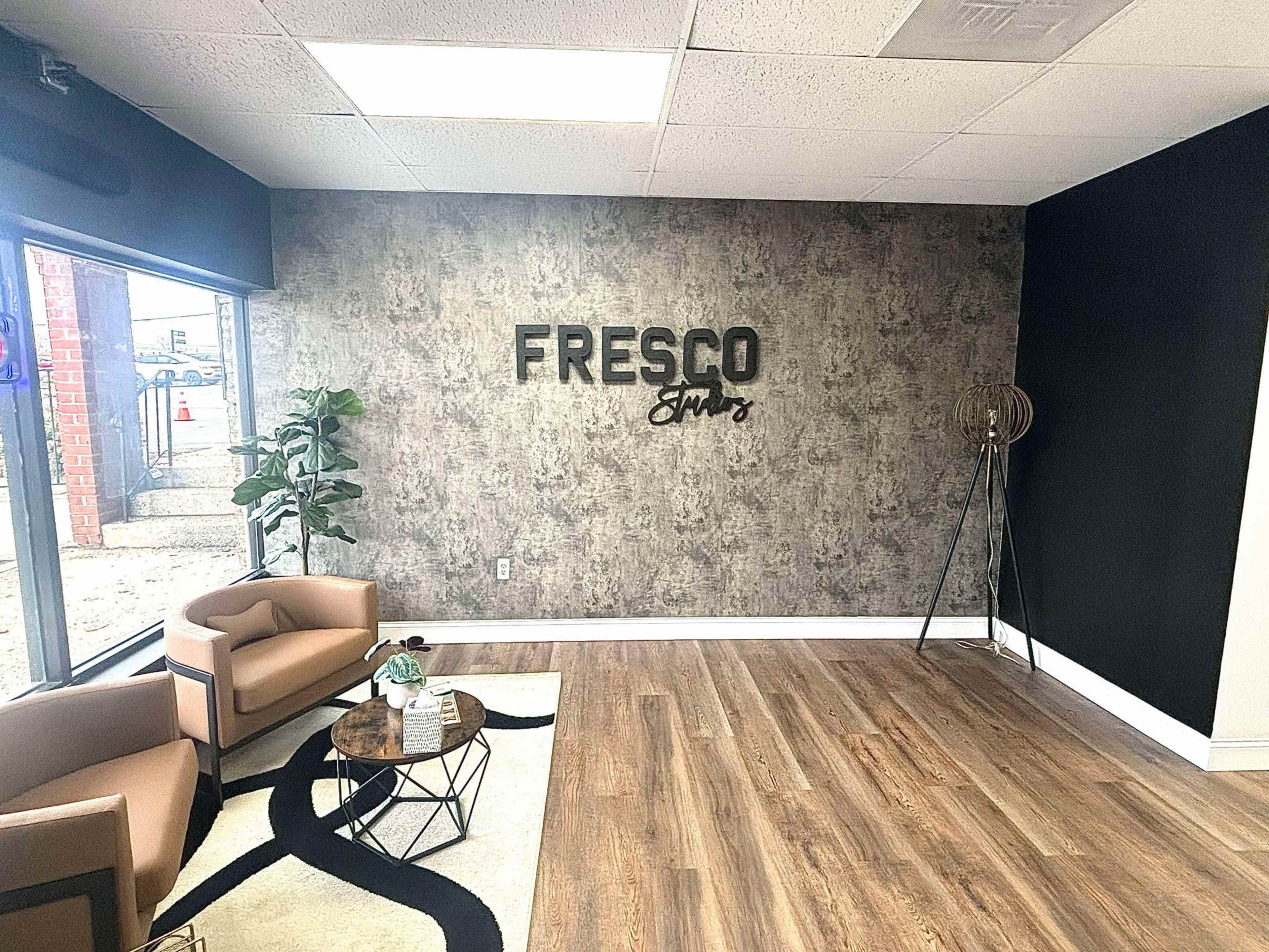 The image shows a modern interior space featuring a brown seating arrangement, a decorative wall with the word "FRESCO", a plant, and a small coffee table on a patterned rug.
