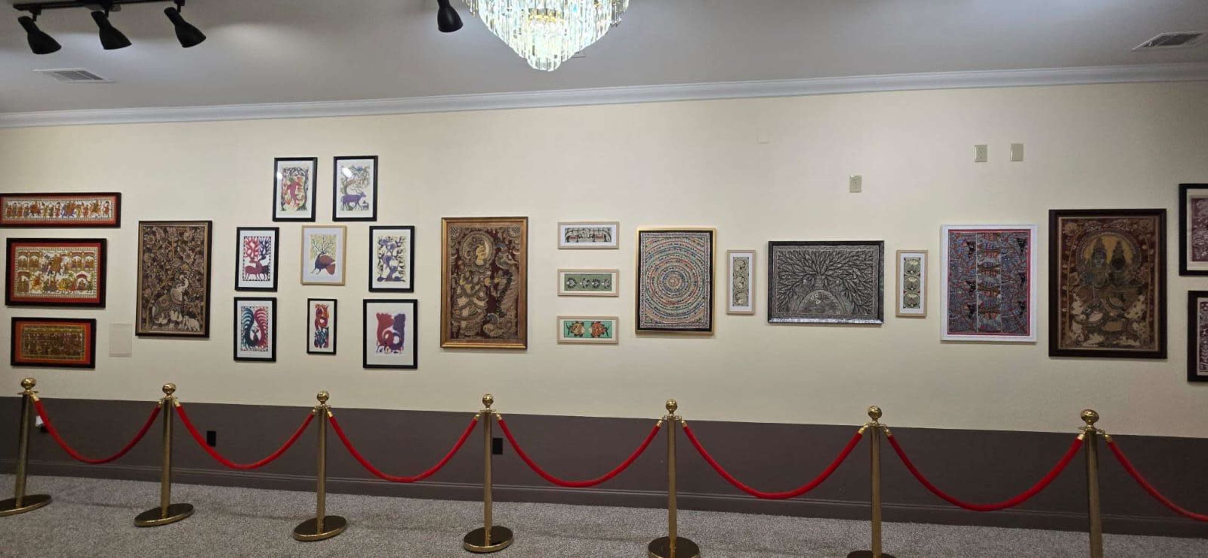 The image shows a gallery wall adorned with various framed artworks, accompanied by a red velvet rope barrier in front.