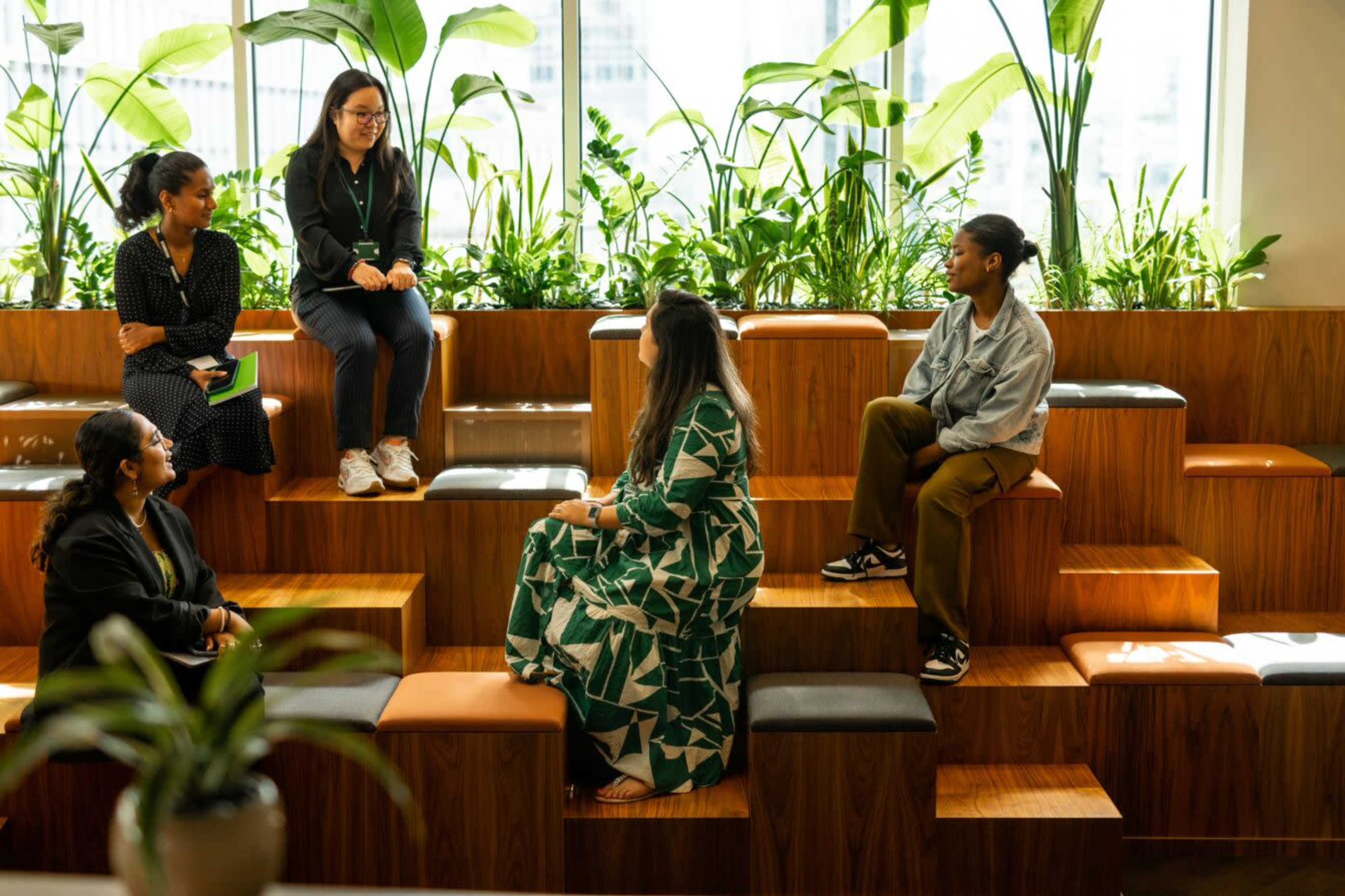 A group of five women are seated on wooden steps in a bright, plant-filled indoor space, engaged in conversation.
