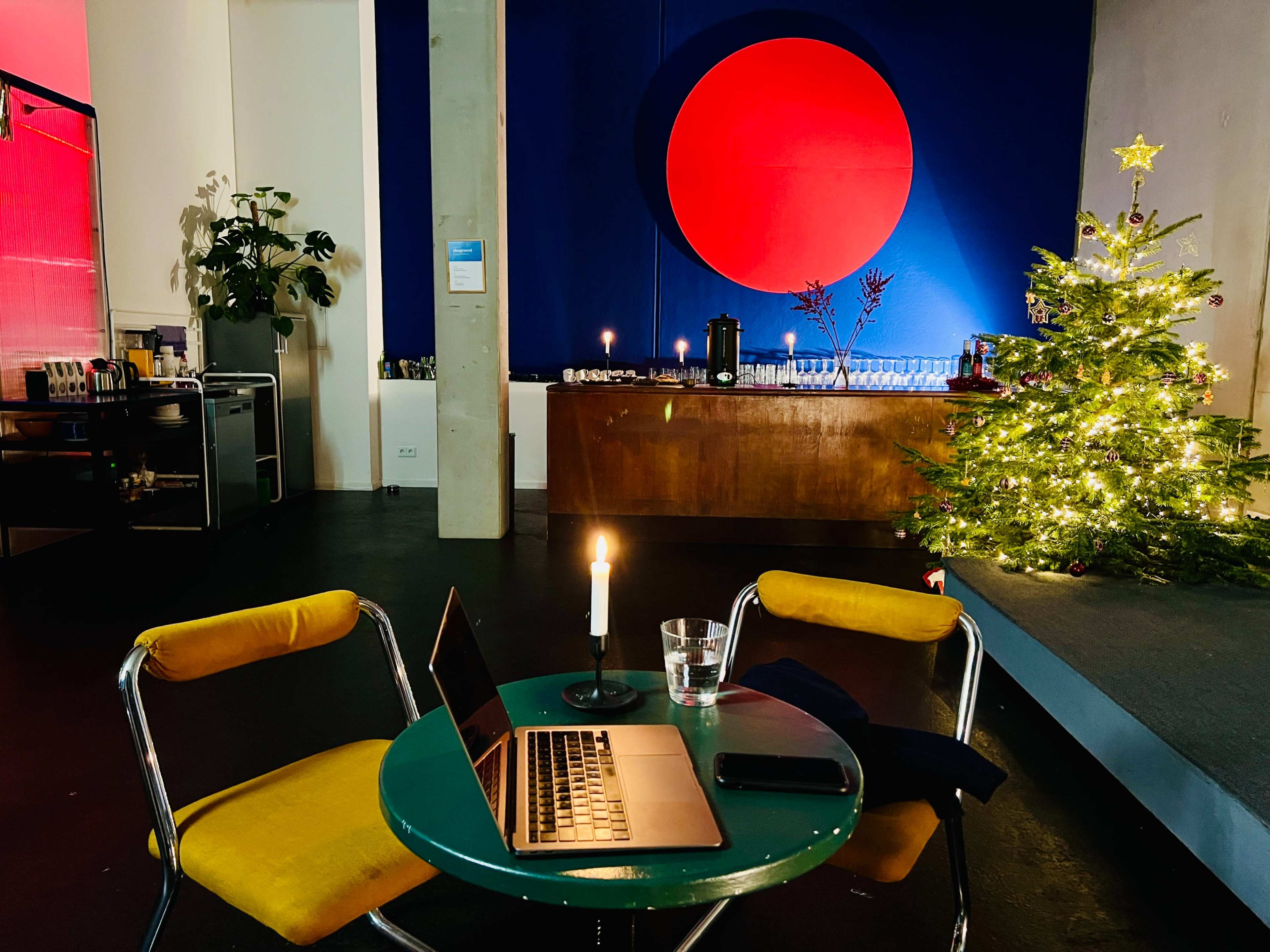 A small green table with a laptop and a candle sits near a decorated Christmas tree in a brightly lit room featuring a large red circle artwork on the wall.