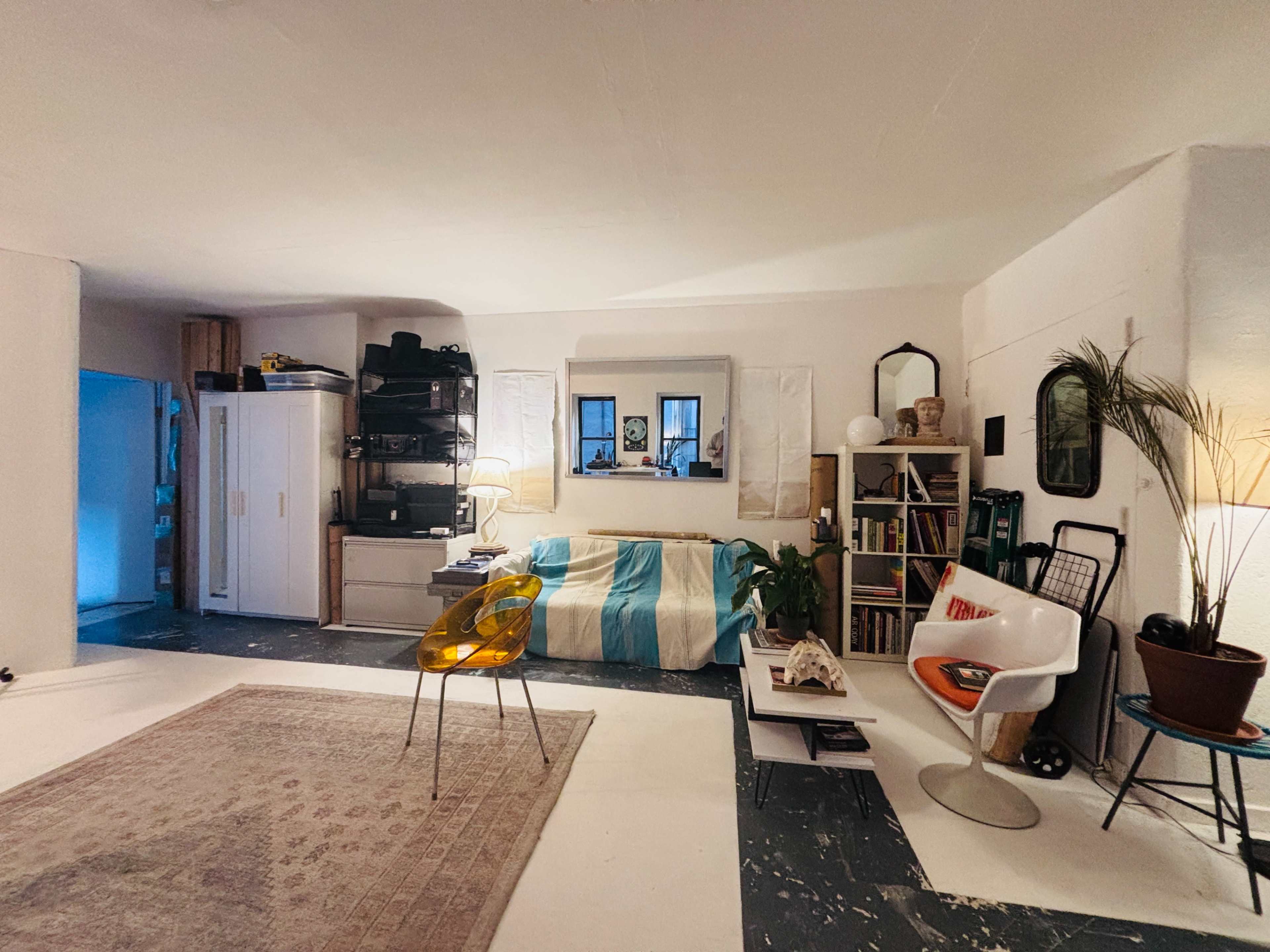 The image shows a cozy living room with a couch, bookshelves, a chair, and a rug, featuring a mixture of furniture and decor in a clean, minimalist style.