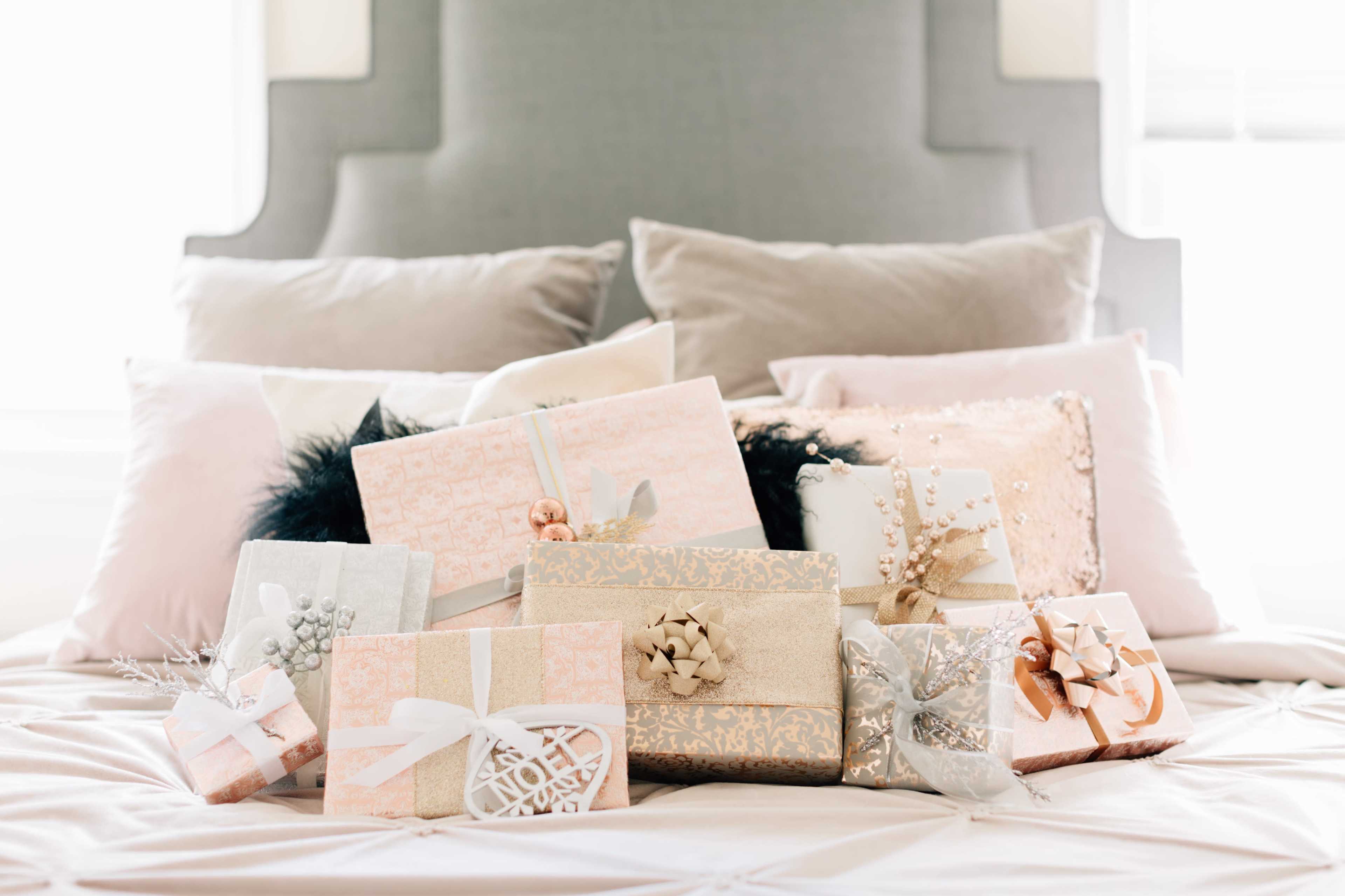 A variety of elegantly wrapped presents are arranged on a bed with plush pillows in a softly lit room.
