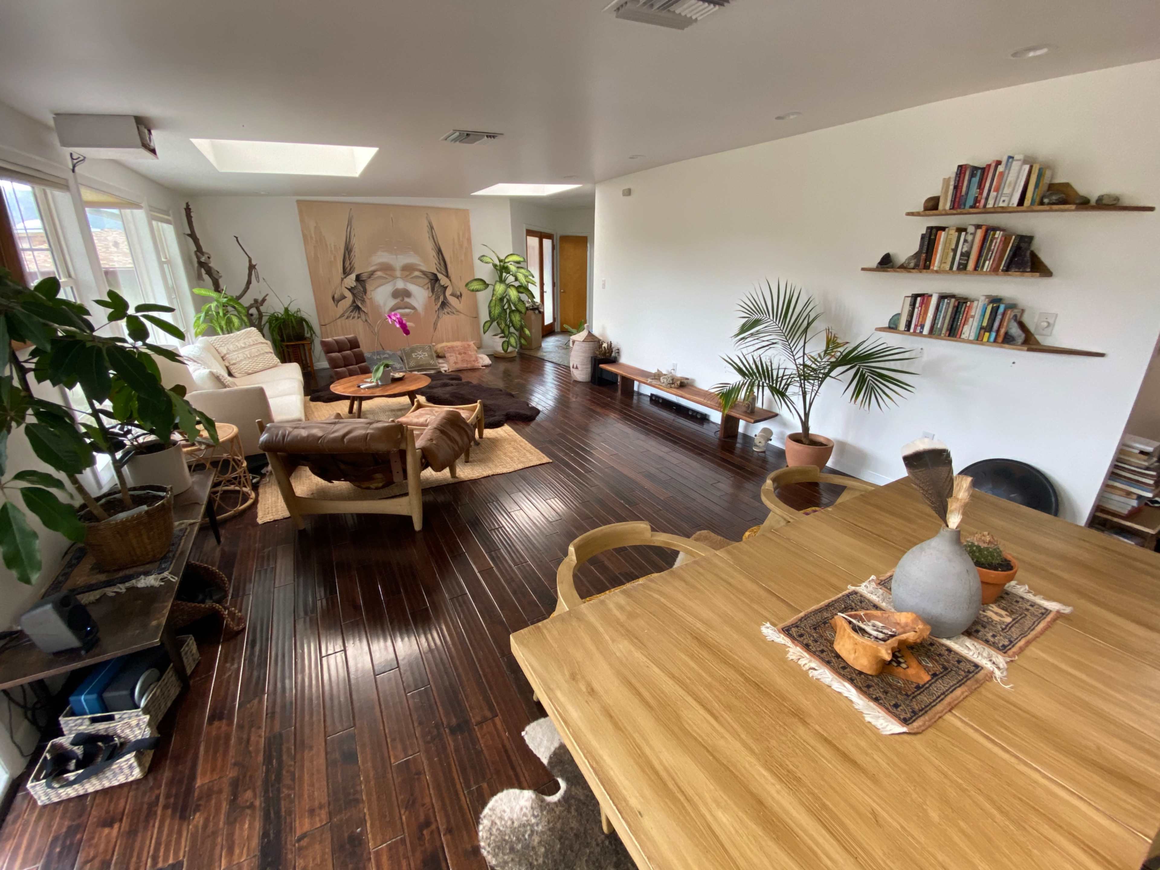 A spacious living room with hardwood floors, a dining table, a sectional sofa, and several plants arranged around a wall featuring a mural.
