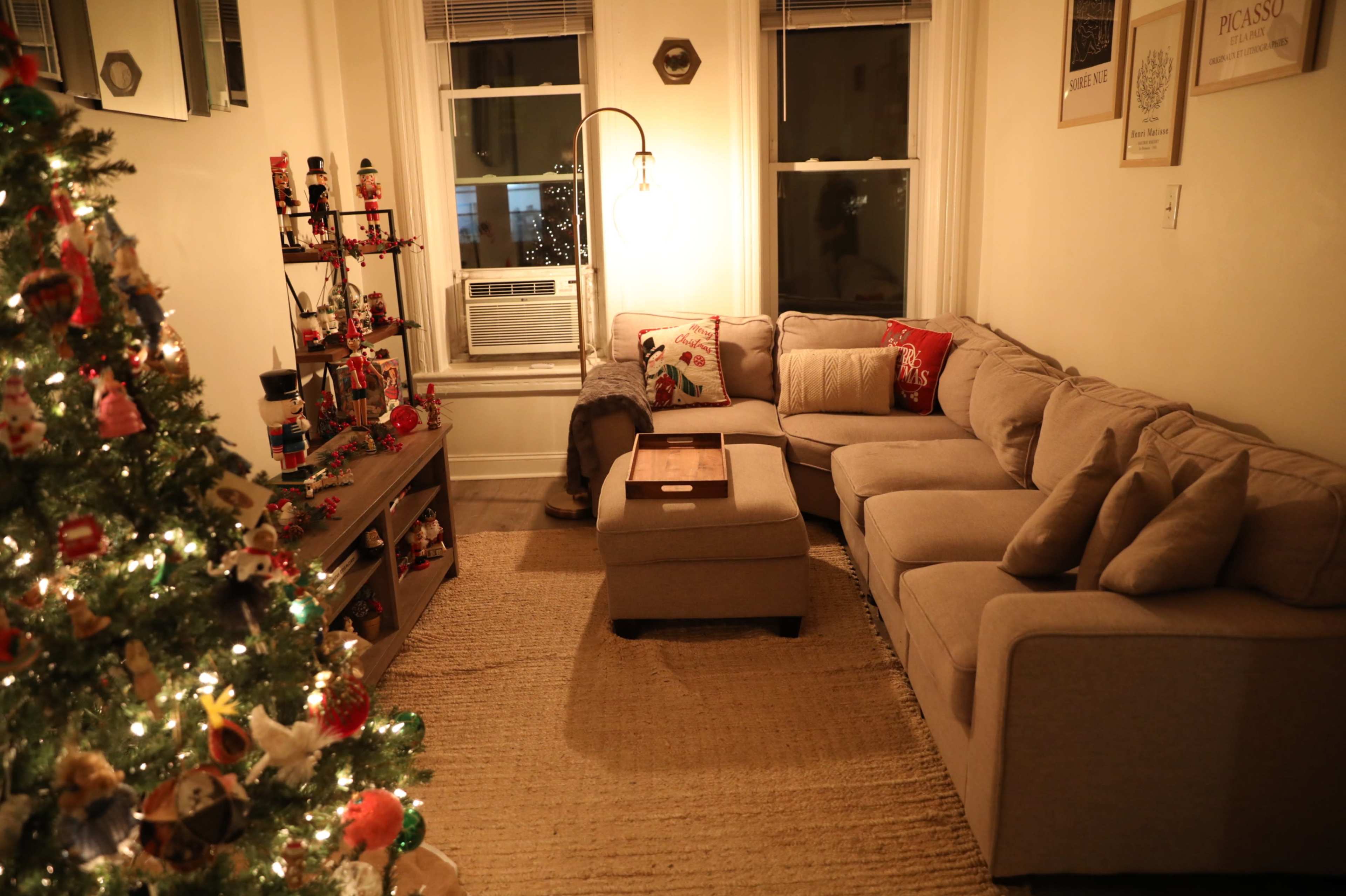 A cozy living room features a sectional sofa, a coffee table, and a Christmas tree adorned with decorations, set against a backdrop of warm lighting and windows.