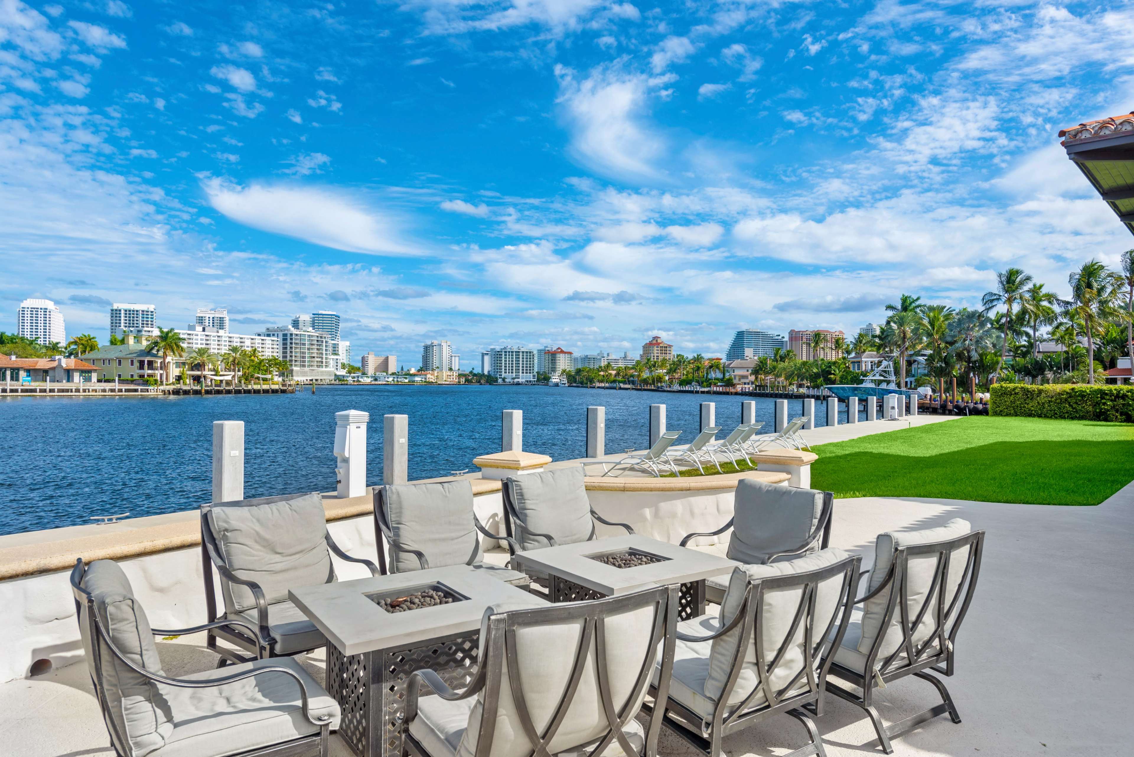 A patio with gray seating surrounds a fire table overlooks a wide waterway with buildings and palm trees in the background under a partly cloudy sky.