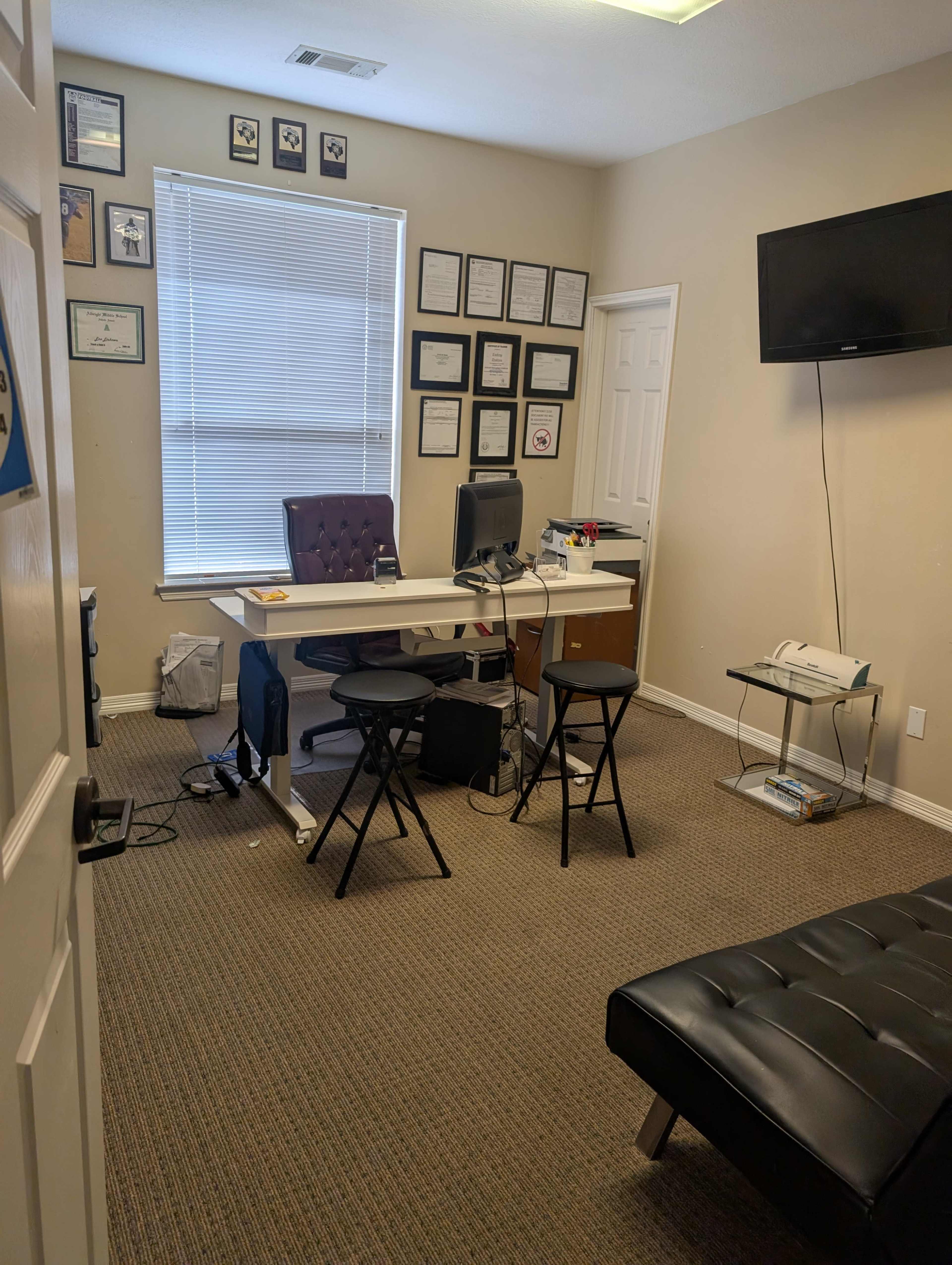 The image shows a small office space with a desk, a computer, a few chairs, and wall-mounted certificates in frames.