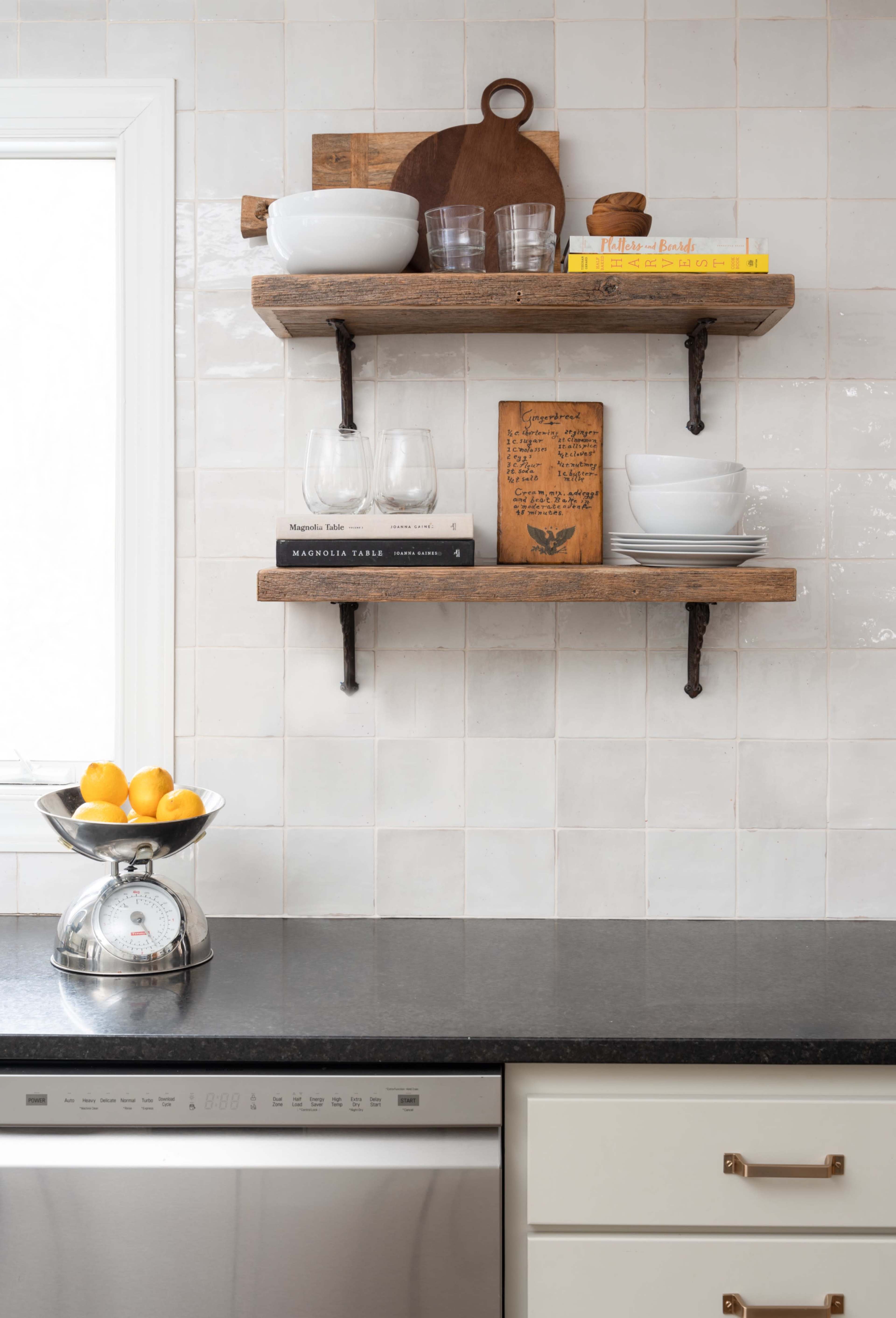 Two wooden shelves with glassware, plates, and cookbooks are mounted on a tiled wall above a black countertop with a scale and a bowl of lemons.