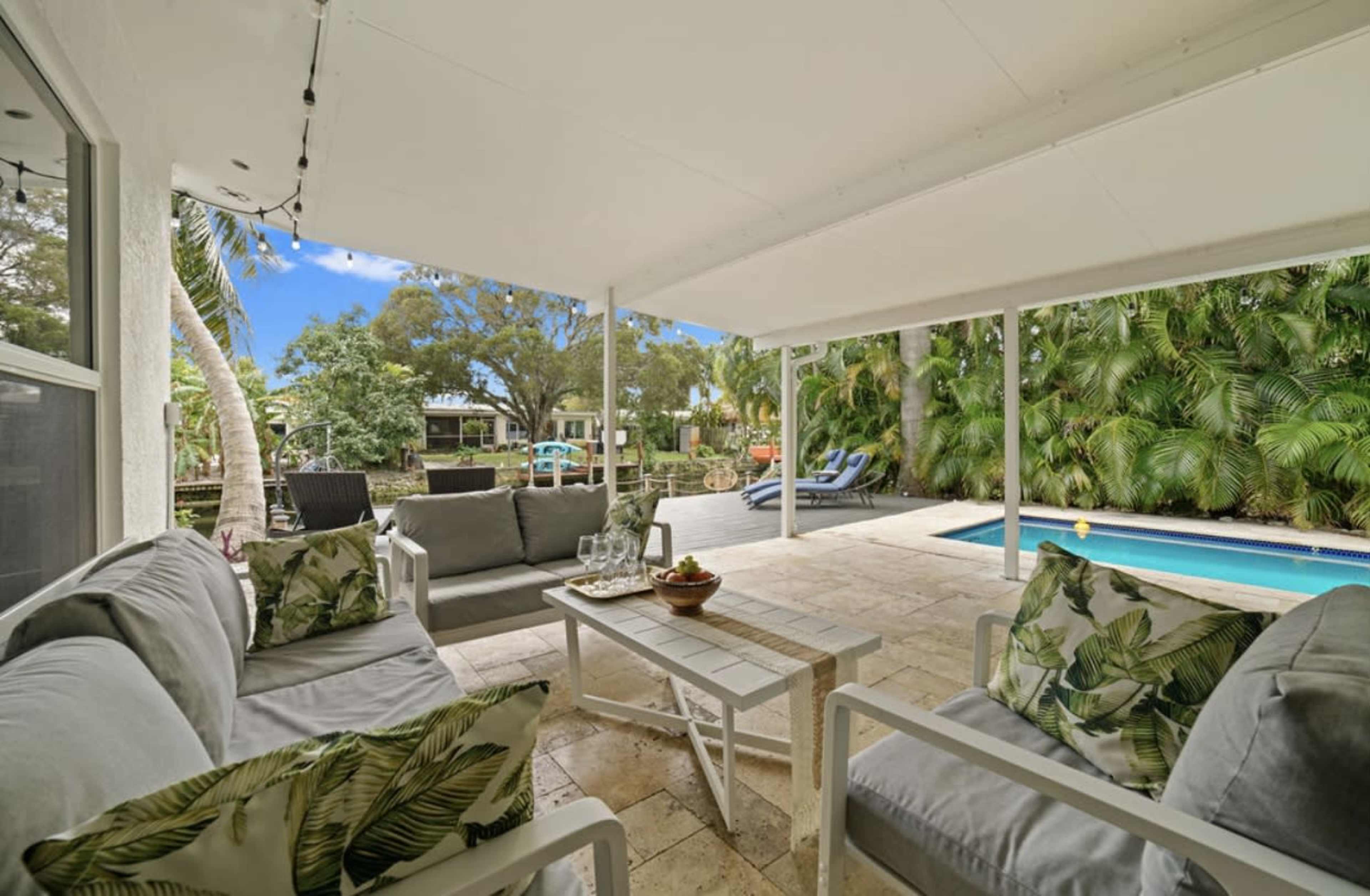 A covered patio area features comfortable seating, a coffee table with snacks, and a view of a pool surrounded by lush greenery.