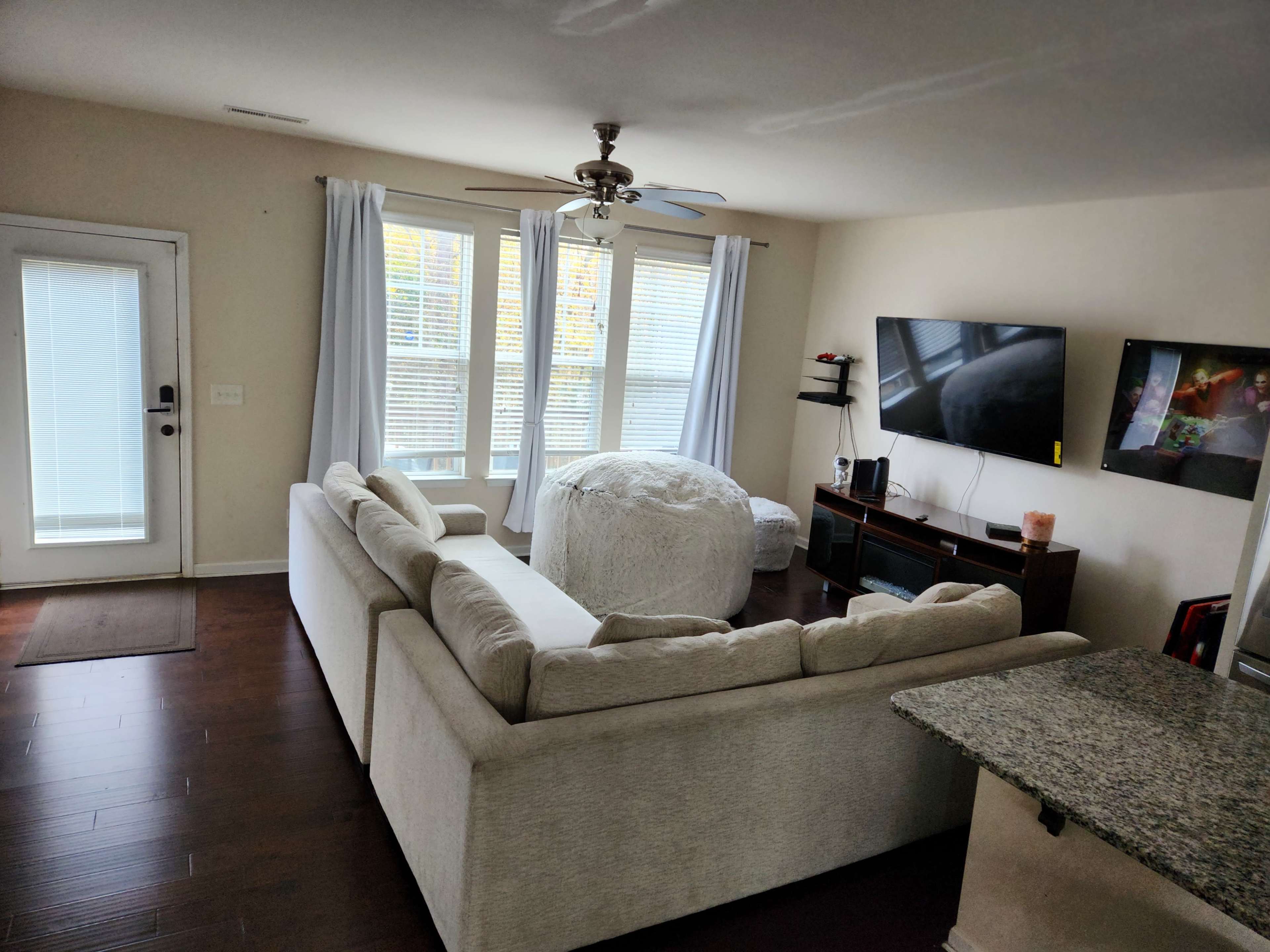 The image shows a living room with a light-colored sectional sofa, a round coffee table, a television mounted on the wall, and large windows with sheer curtains.
