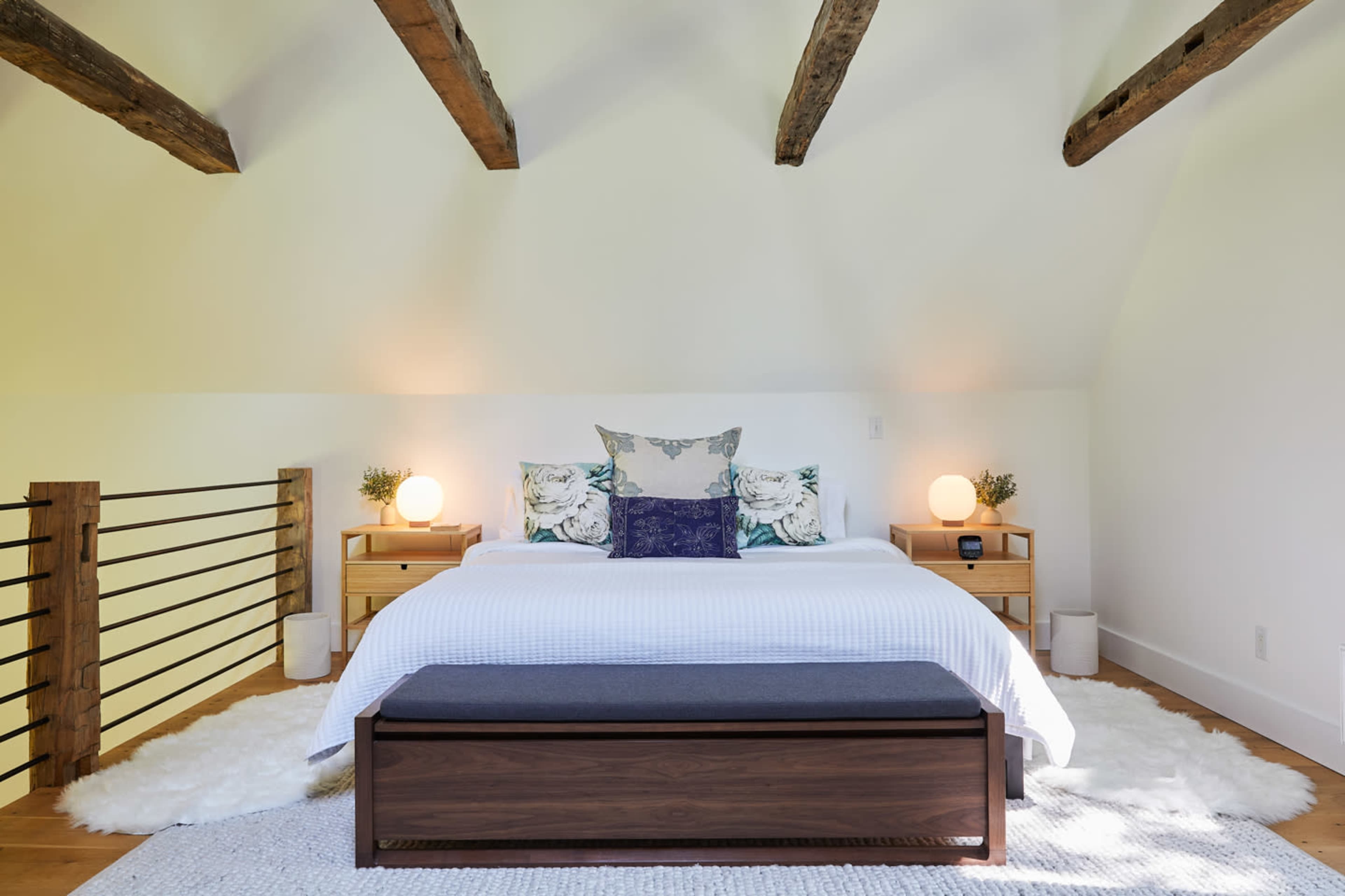 A well-lit bedroom features a bed with decorative pillows, two bedside tables with lamps, and a bench at the foot on a soft rug.
