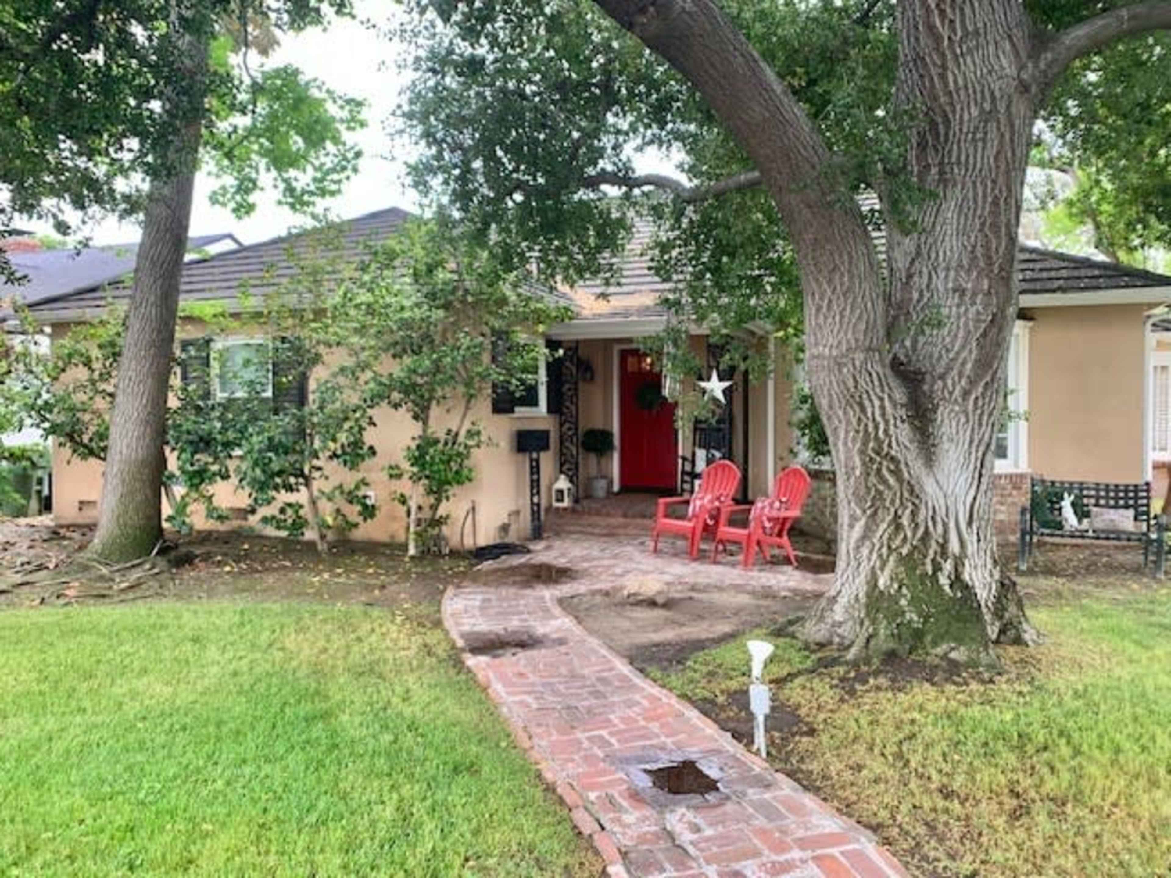 A brick pathway leads to the entrance of a single-story house framed by large trees and red seating on the porch.