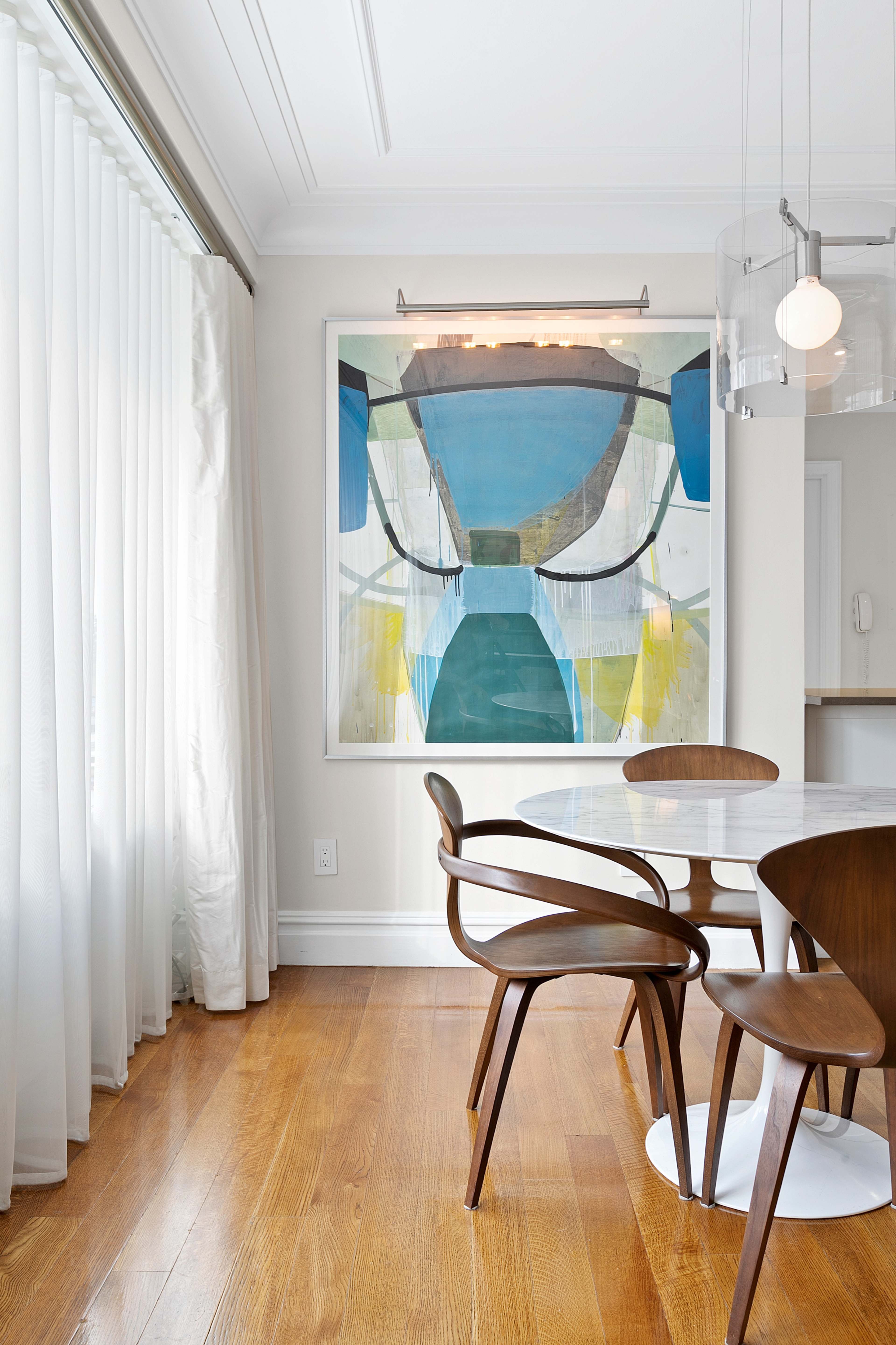A dining area features a round white table surrounded by wooden chairs, with large windows dressed in sheer curtains and a colorful abstract painting on the wall.