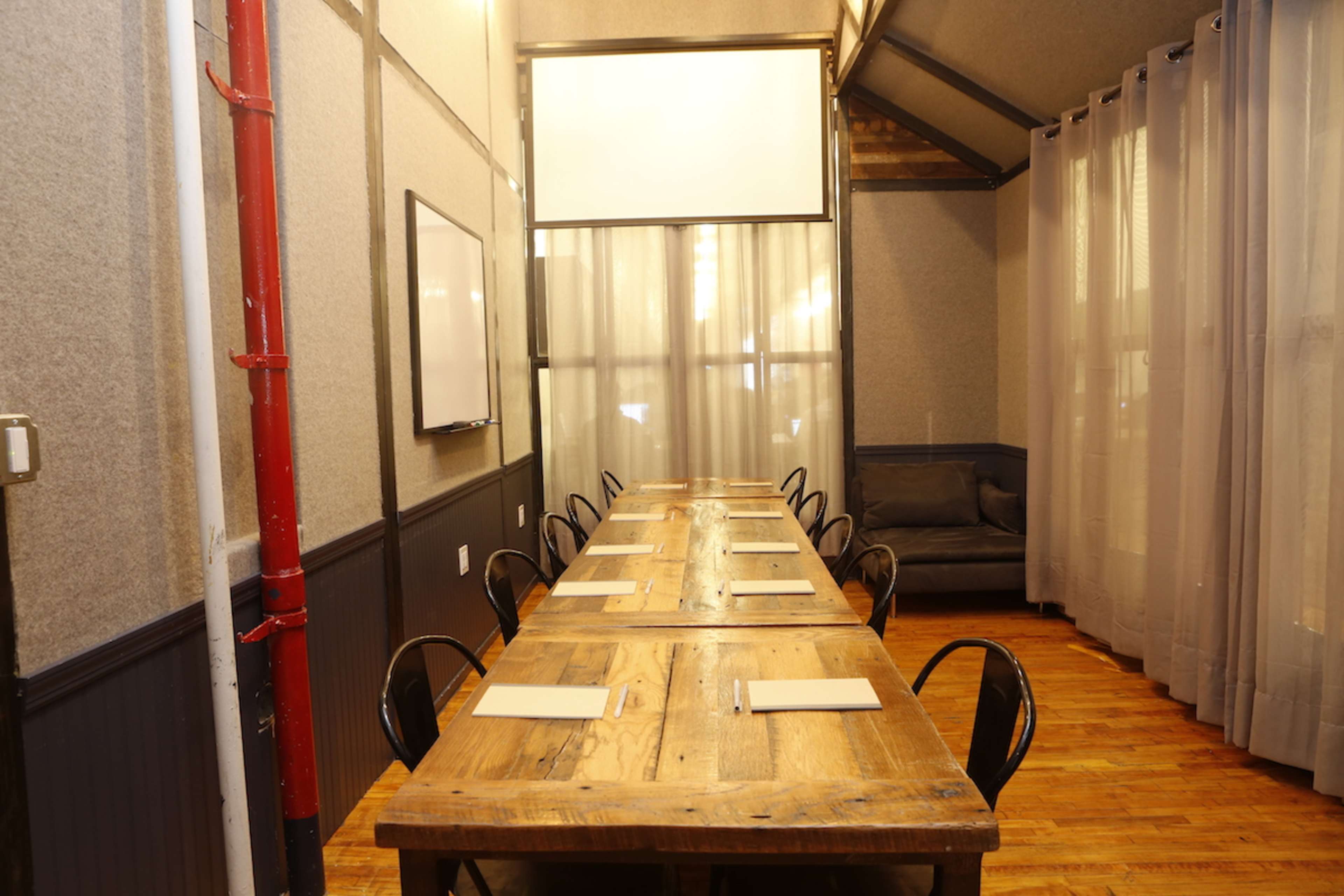 A long wooden table is set for a meeting in a room with gray walls and large windows covered by sheer curtains.