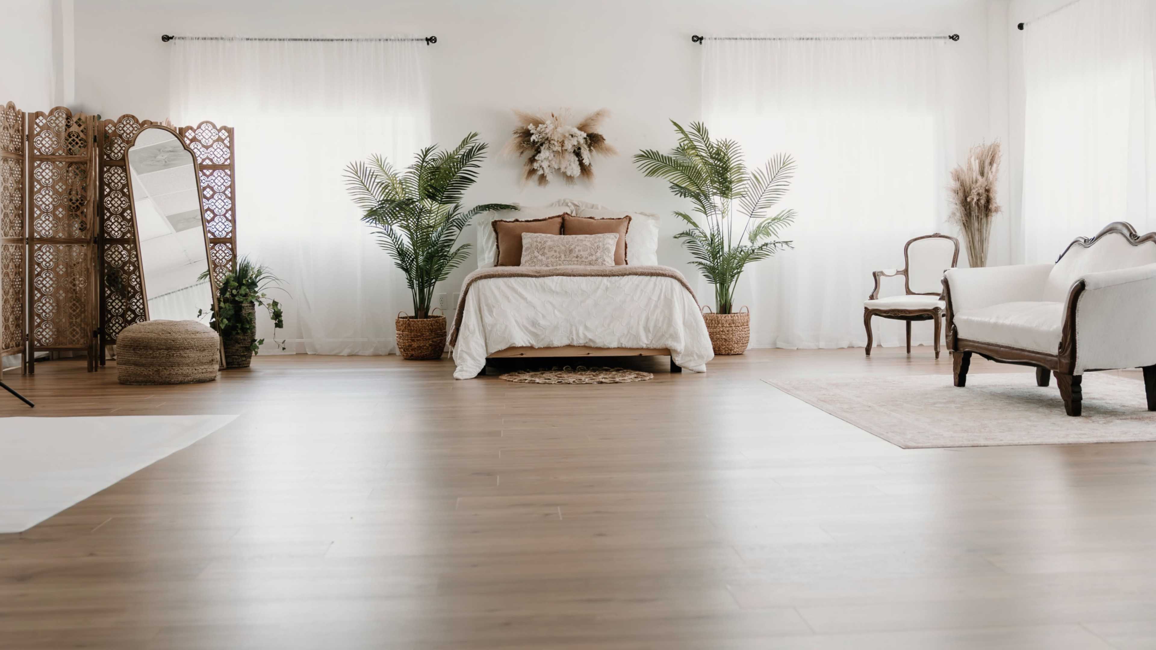 The scene depicts a bright bedroom with a bed adorned in white linens, surrounded by potted plants, a wooden chair, and a decorative screen.