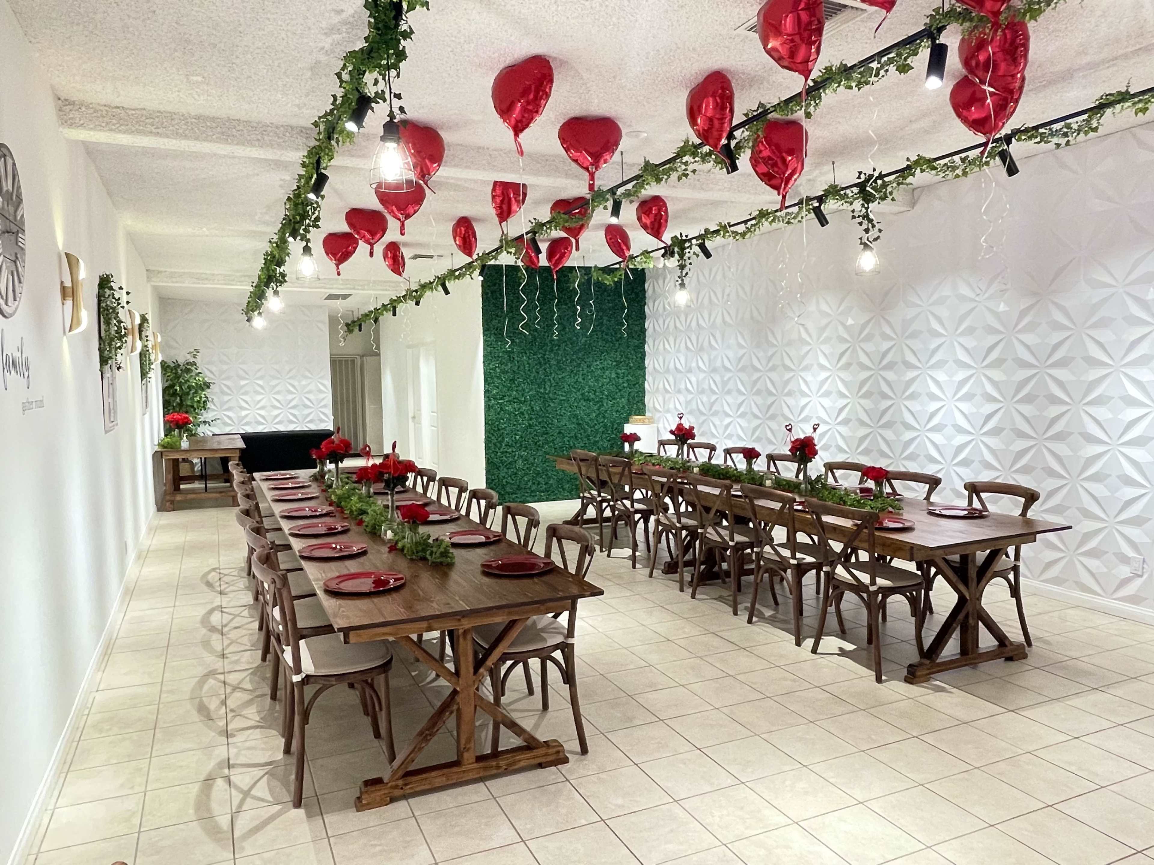 A spacious dining area features wooden tables set with red plates and floral centerpieces, adorned with red heart-shaped balloons and greenery hanging from the ceiling.
